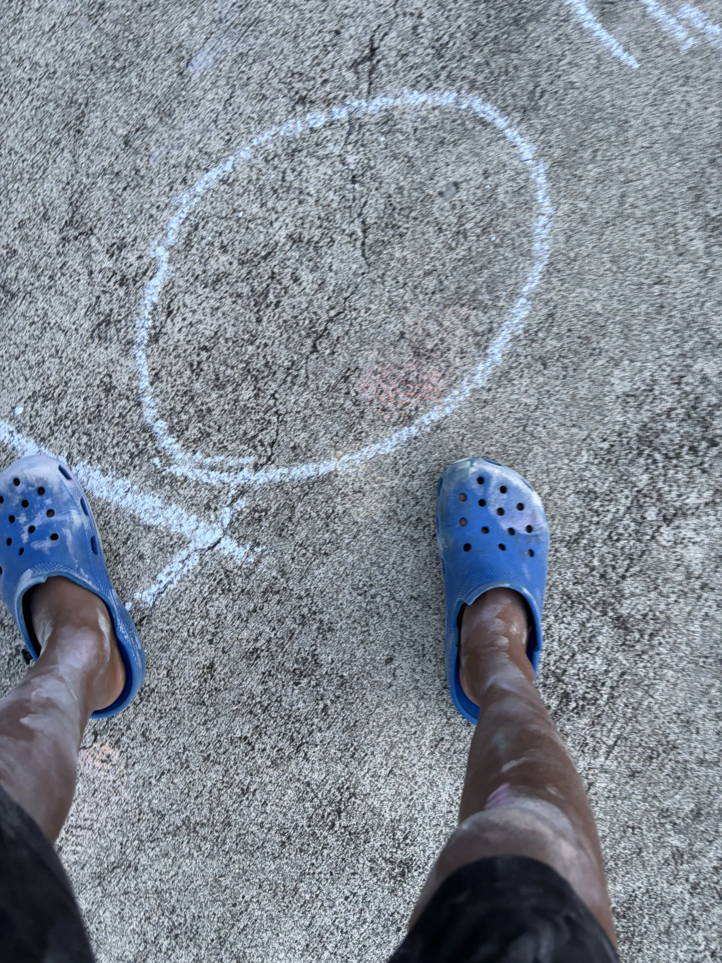 Person standing on a textured concrete surface wearing blue Crocs, with a chalk-drawn circle on the ground.