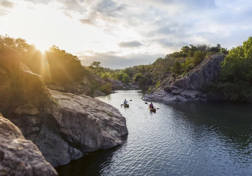 Two people kayaking on a calm river surrounded by rocky cliffs and green trees, with the sun setting behind the landscape.