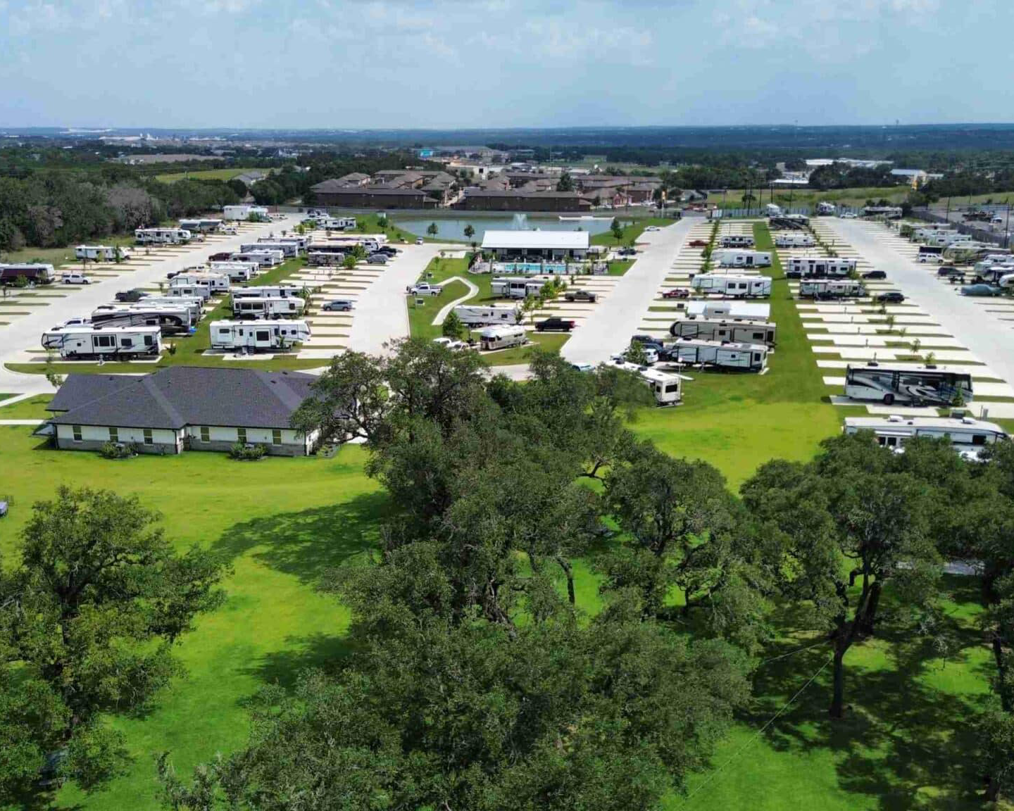 A large RV park with numerous RVs parked in designated spaces, surrounded by lush green trees and grass, with a building and pond nearby.