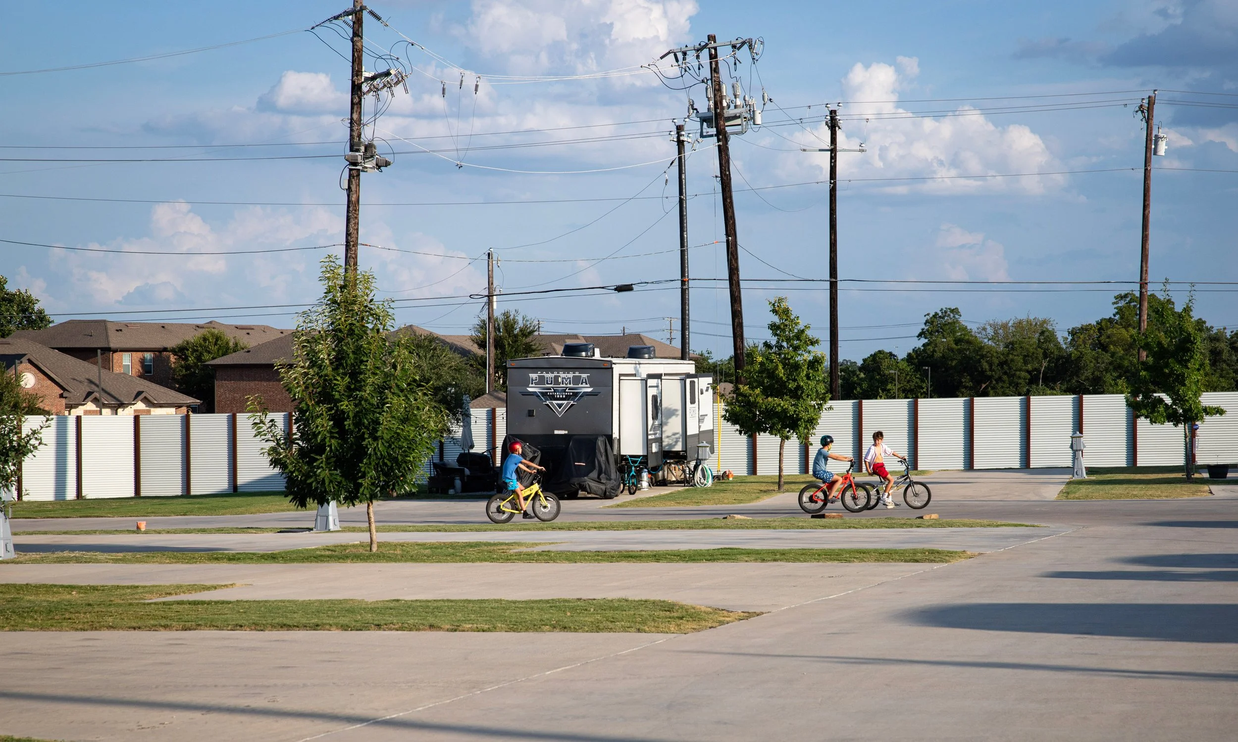 Children riding bicycles in a park with trees, a white fence, and houses in the background. Utility poles and power lines are visible in the sky.