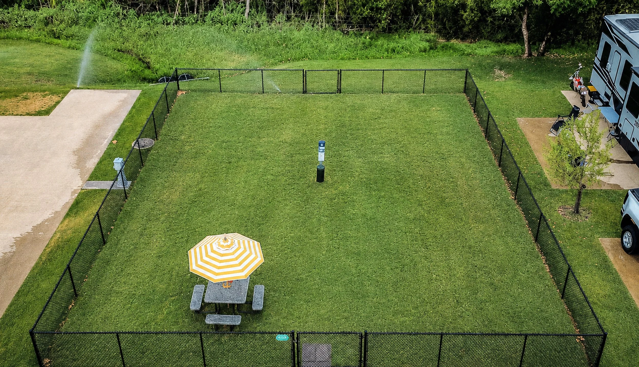 A fenced-in backyard with a grassy area, a picnic table with an umbrella, and a trailer adjacent to the yard. There are sprinklers watering the grass and a small tree in the yard.