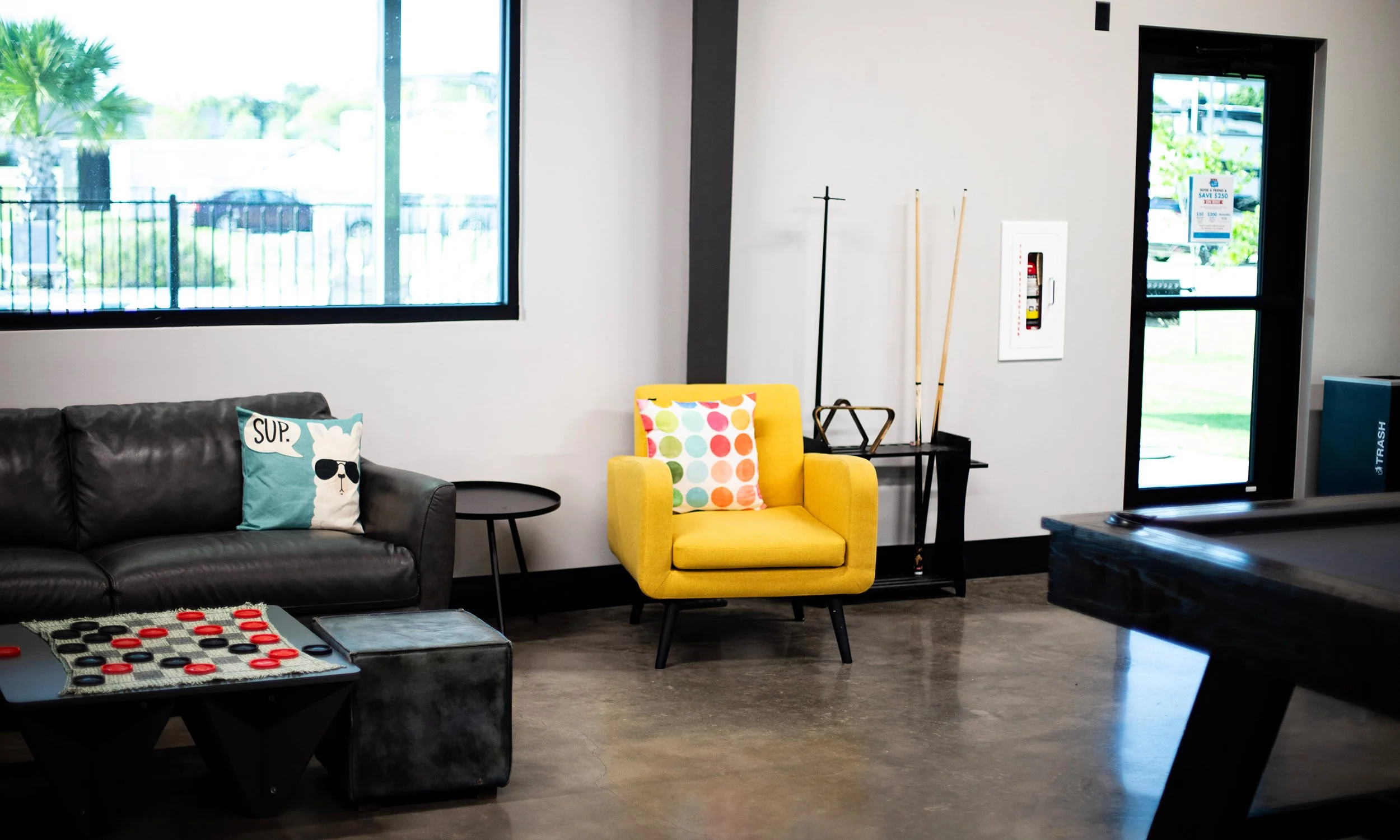 Interior of a lounge or waiting area with a black leather couch, a yellow armchair with a colorful polka dot pillow, a small round black side table, a large window, and a door with a sign outside. There is a fire extinguisher in a white box on the wa
