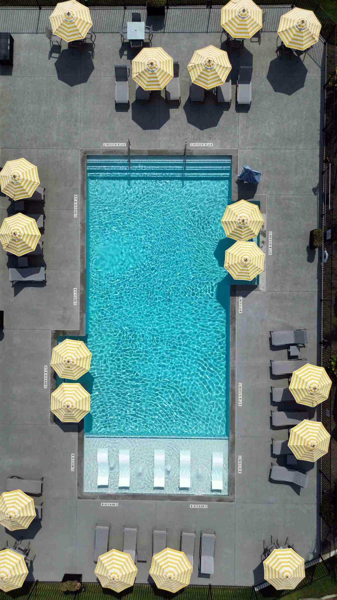 An aerial view of a swimming pool surrounded by yellow and white striped umbrellas and lounge chairs on a concrete deck.