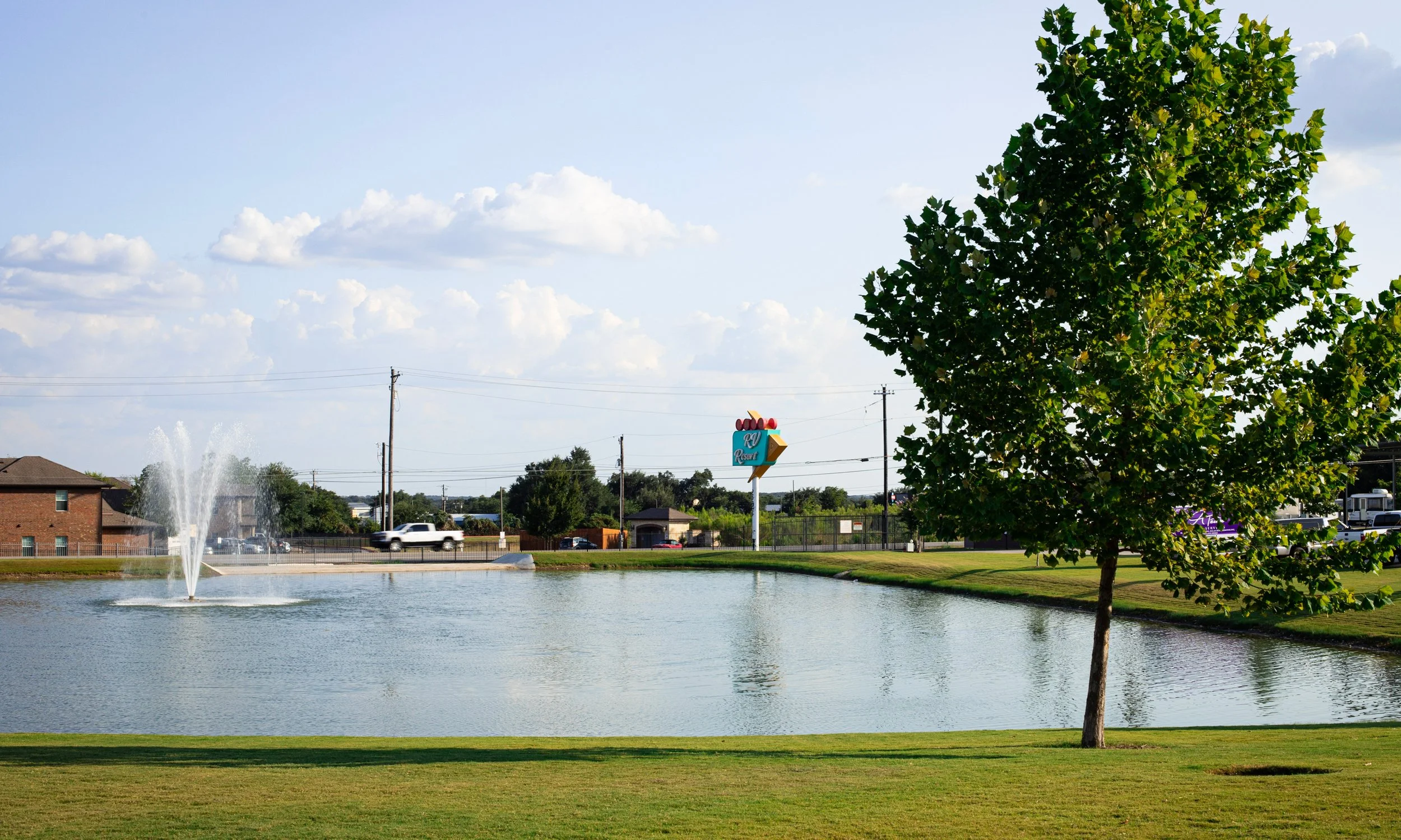 A park with a pond, fountain, large tree, grassy area, and a road with cars. There's a sign for RW Resort in the background.