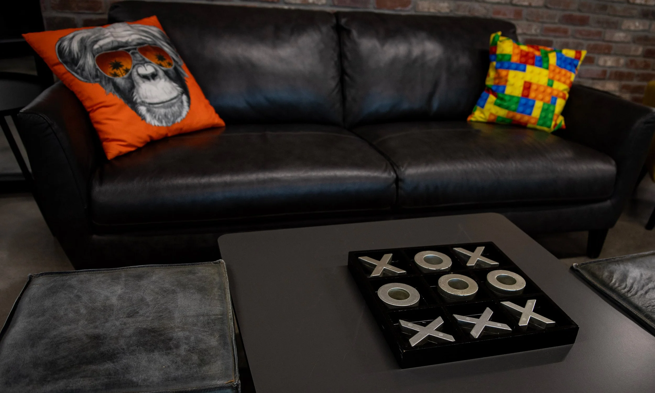 A black leather sofa with two colorful pillows, one featuring a skull with sunglasses and the other with a mosaic pattern, in a room with exposed brick wall and a coffee table with a tic-tac-toe game on it.