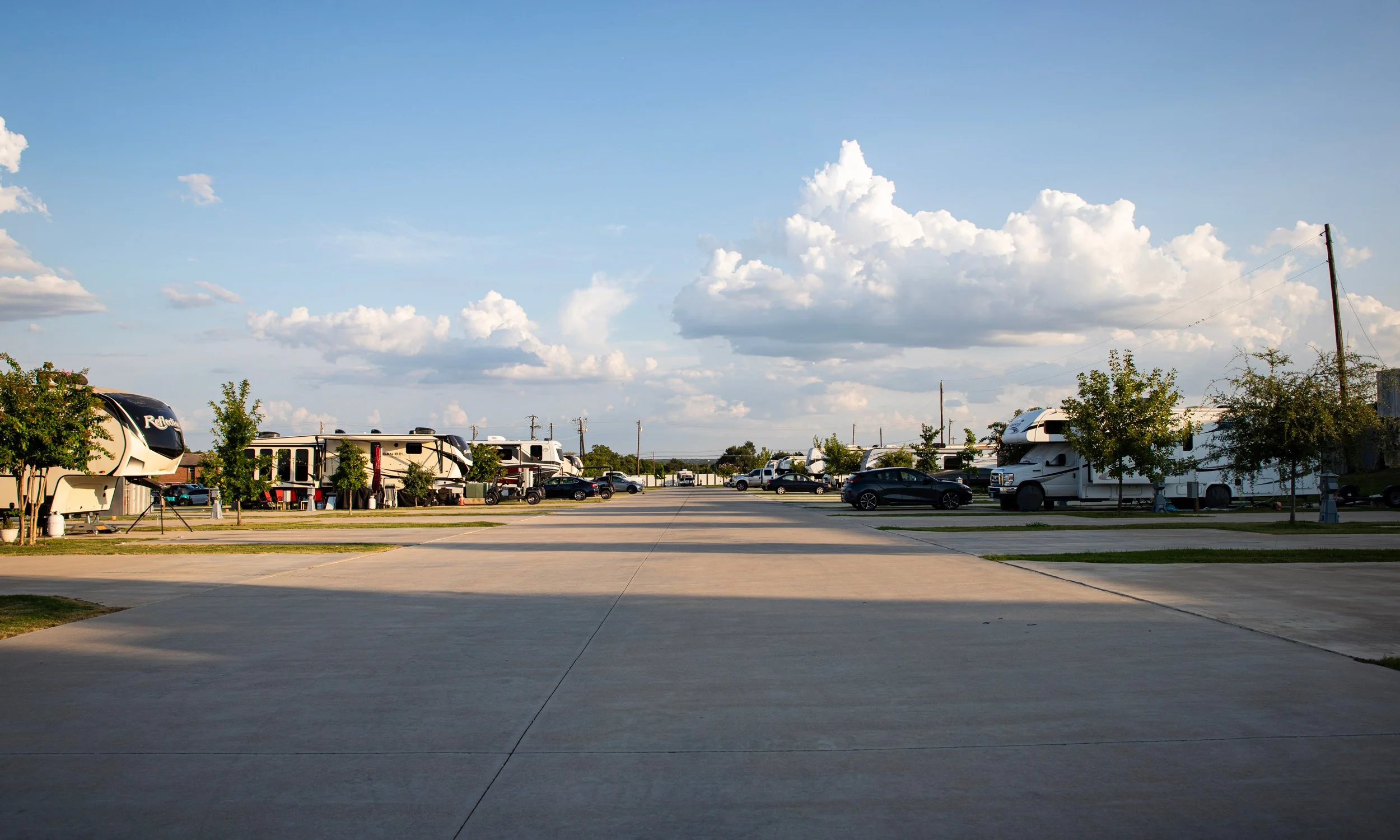 A parking lot with several RVs and cars parked, with some trees and utility poles in the background under a partly cloudy sky.