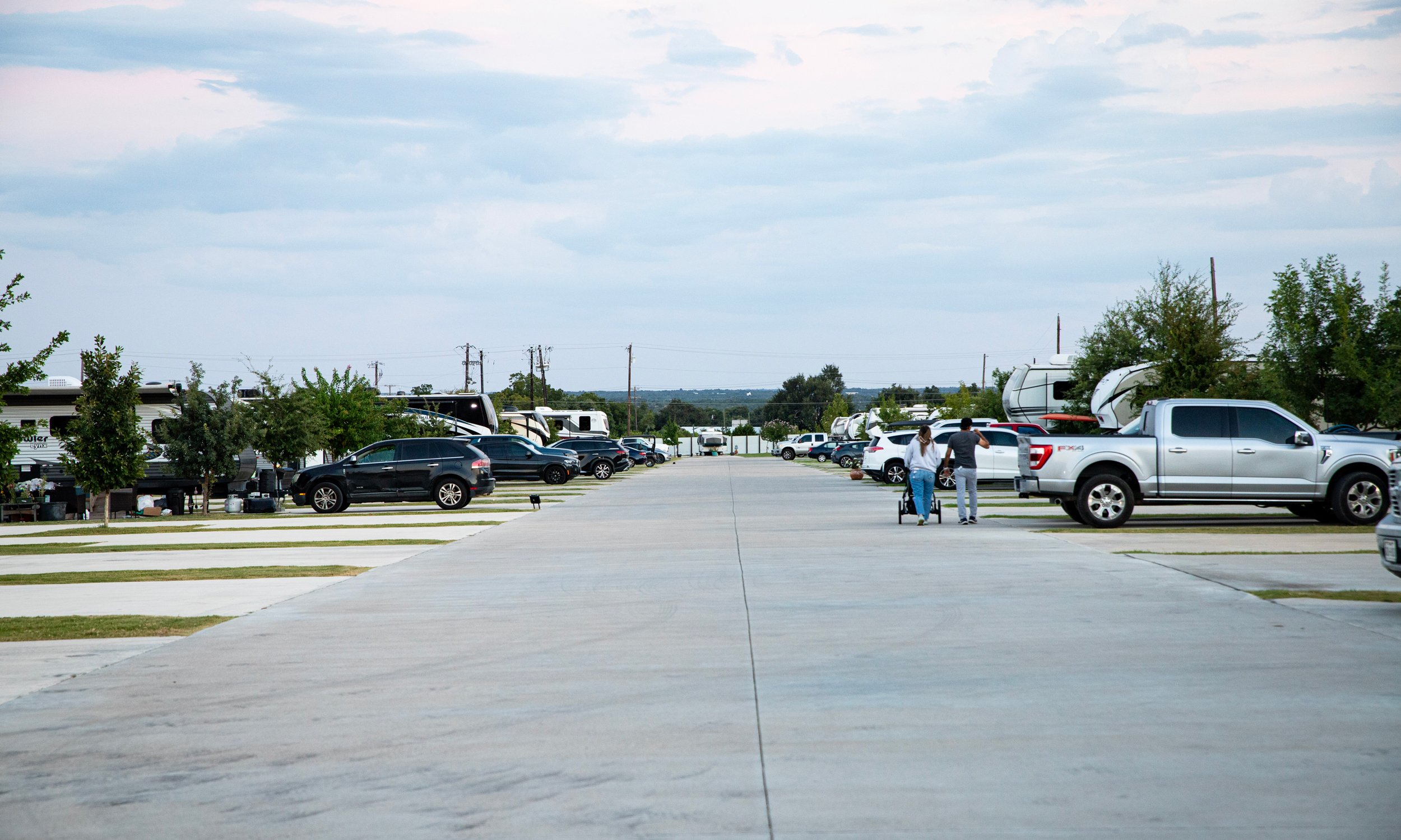 A parking lot with various cars and RVs, a couple walking with shopping carts, and trees along the sidewalk under a partly cloudy sky.