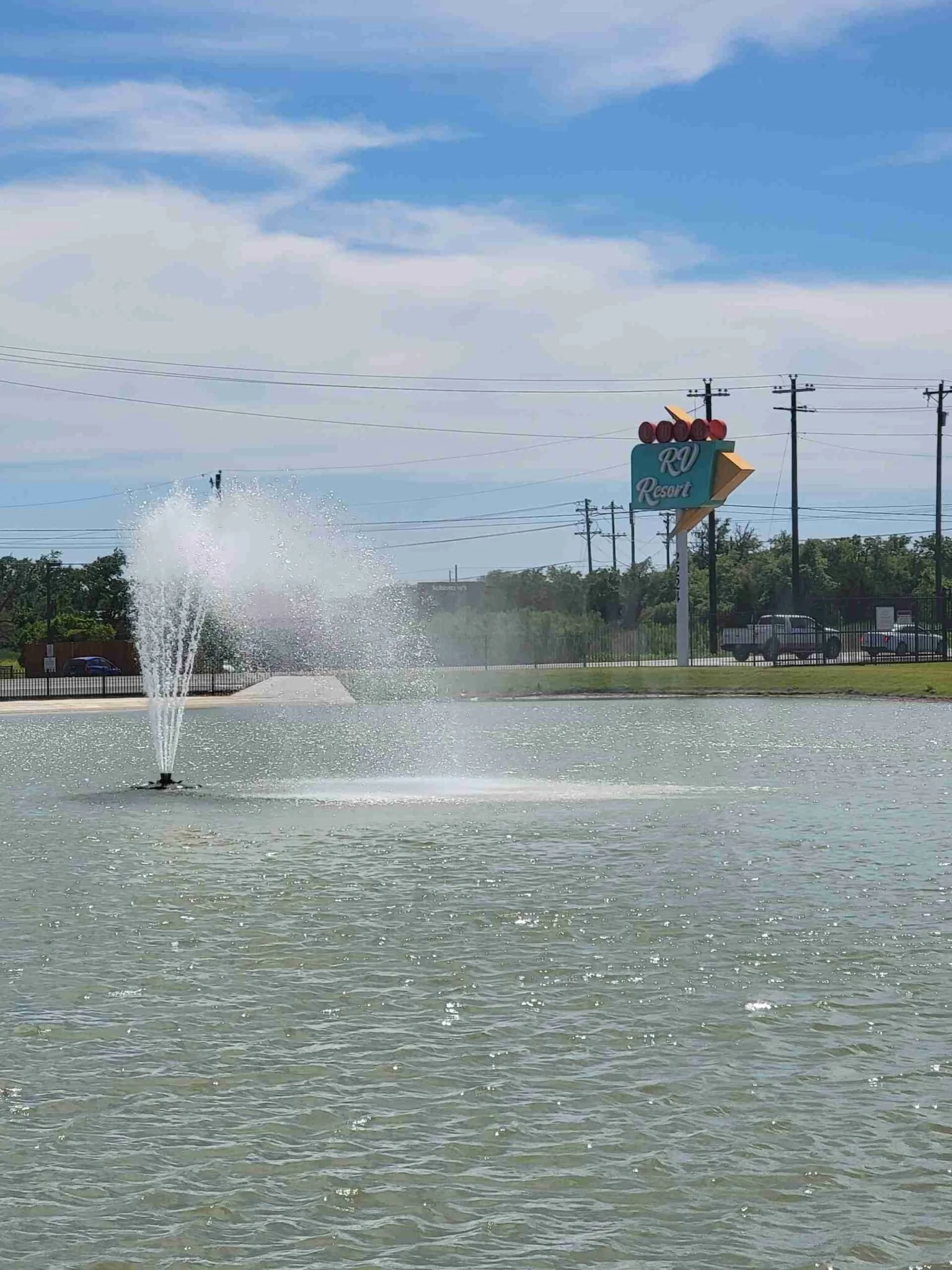 A pond or lake with a fountain in the foreground, water spraying upwards, and a parking lot with cars and a sign for RV Resort in the background