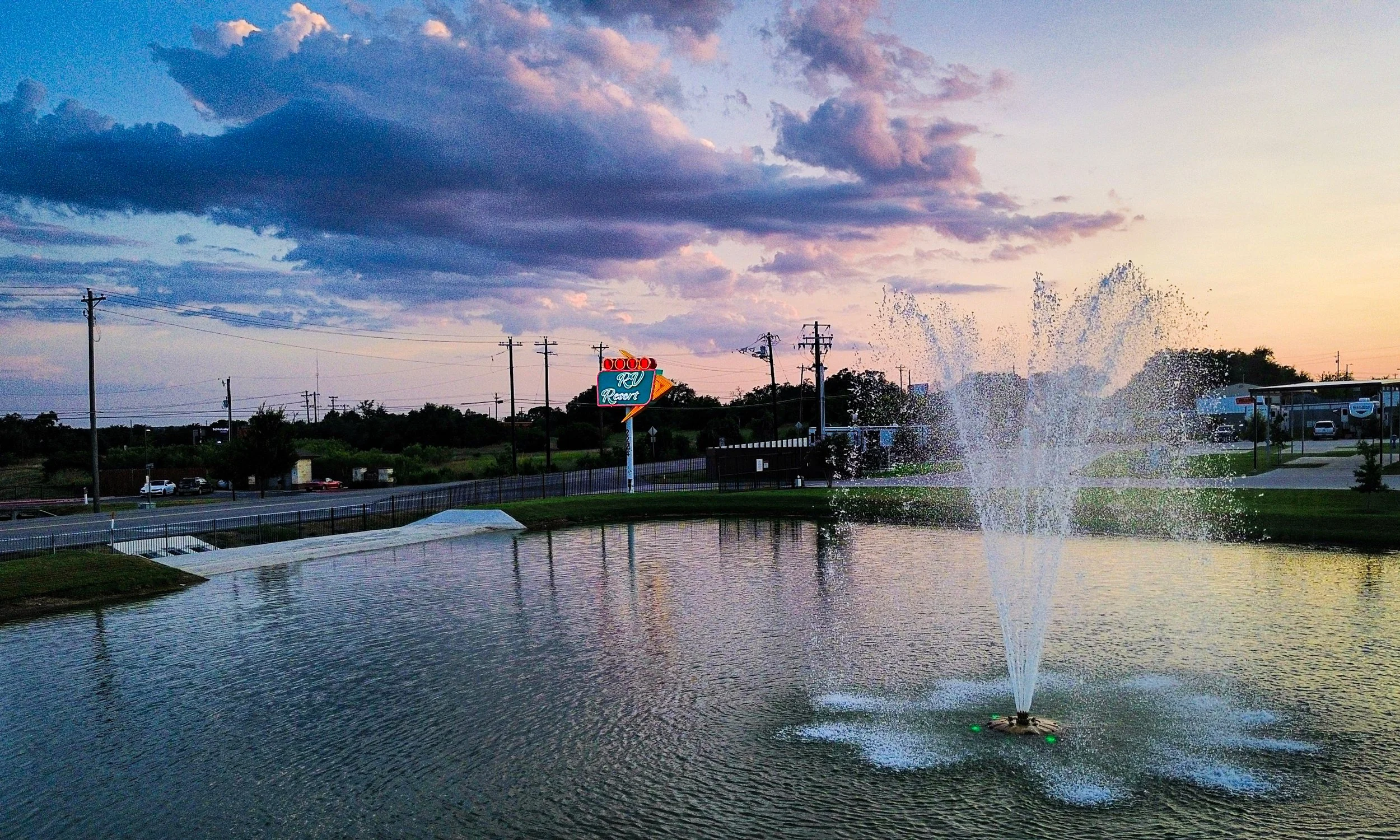 A pond with a fountain spraying water, a road with cars, and a sunset sky with clouds. Visible signage for RV Resort.
