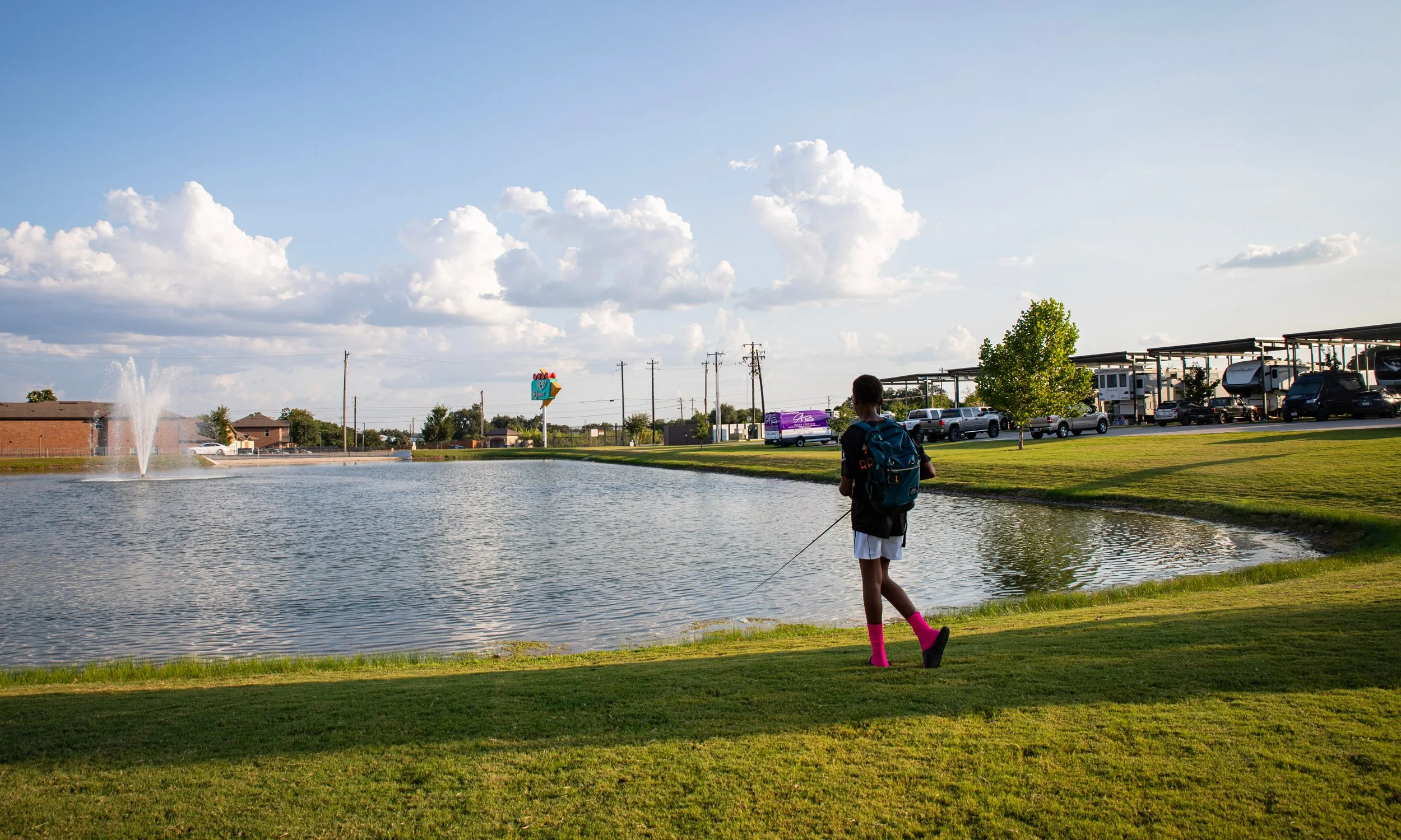 A boy in pink socks fishing by a pond with a fountain, recent clouds, and a parking lot with cars and a RV park in the background.