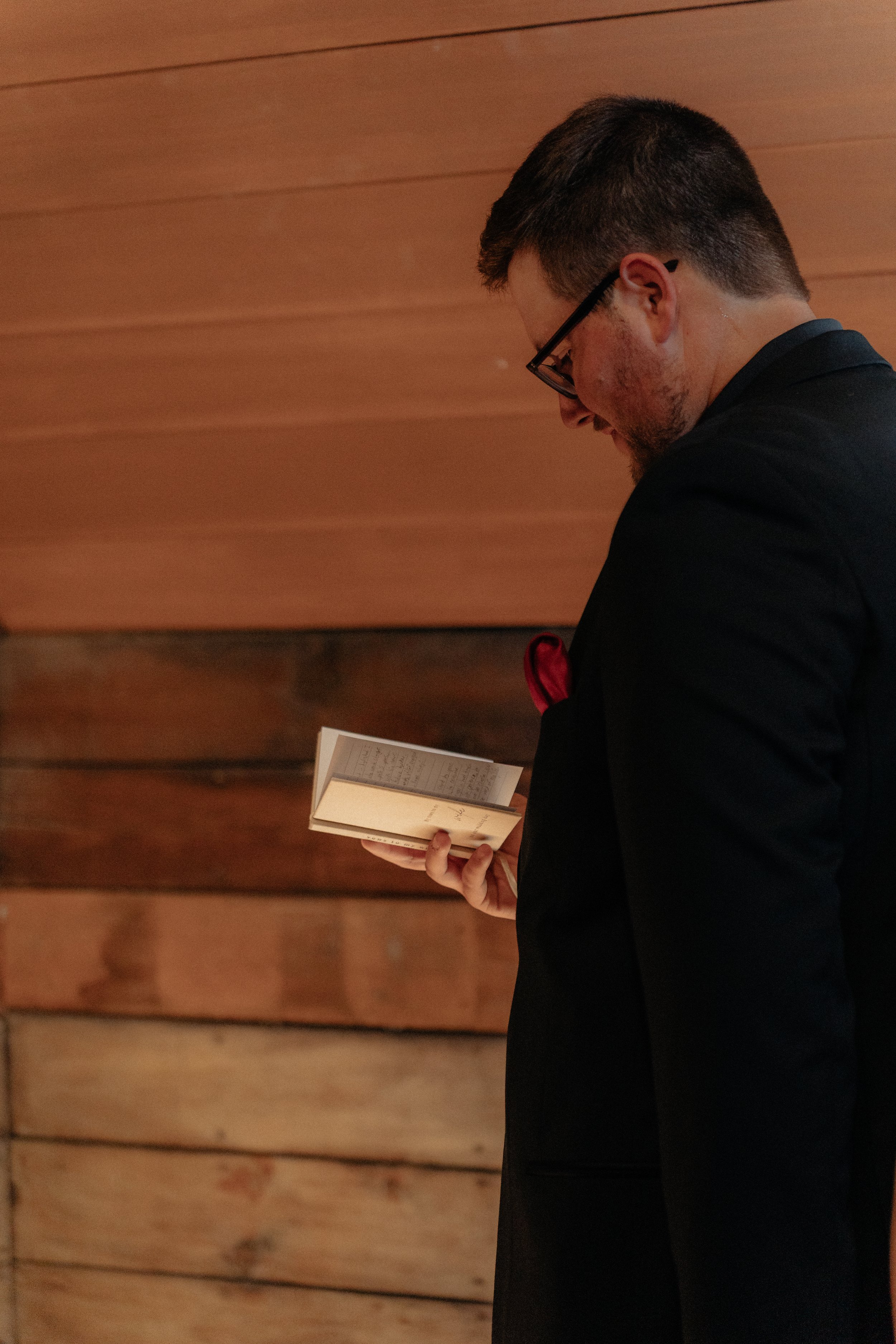 A man with short hair, glasses, and a beard reading a book, wearing a dark suit and a red pocket square, standing against a wooden wall.