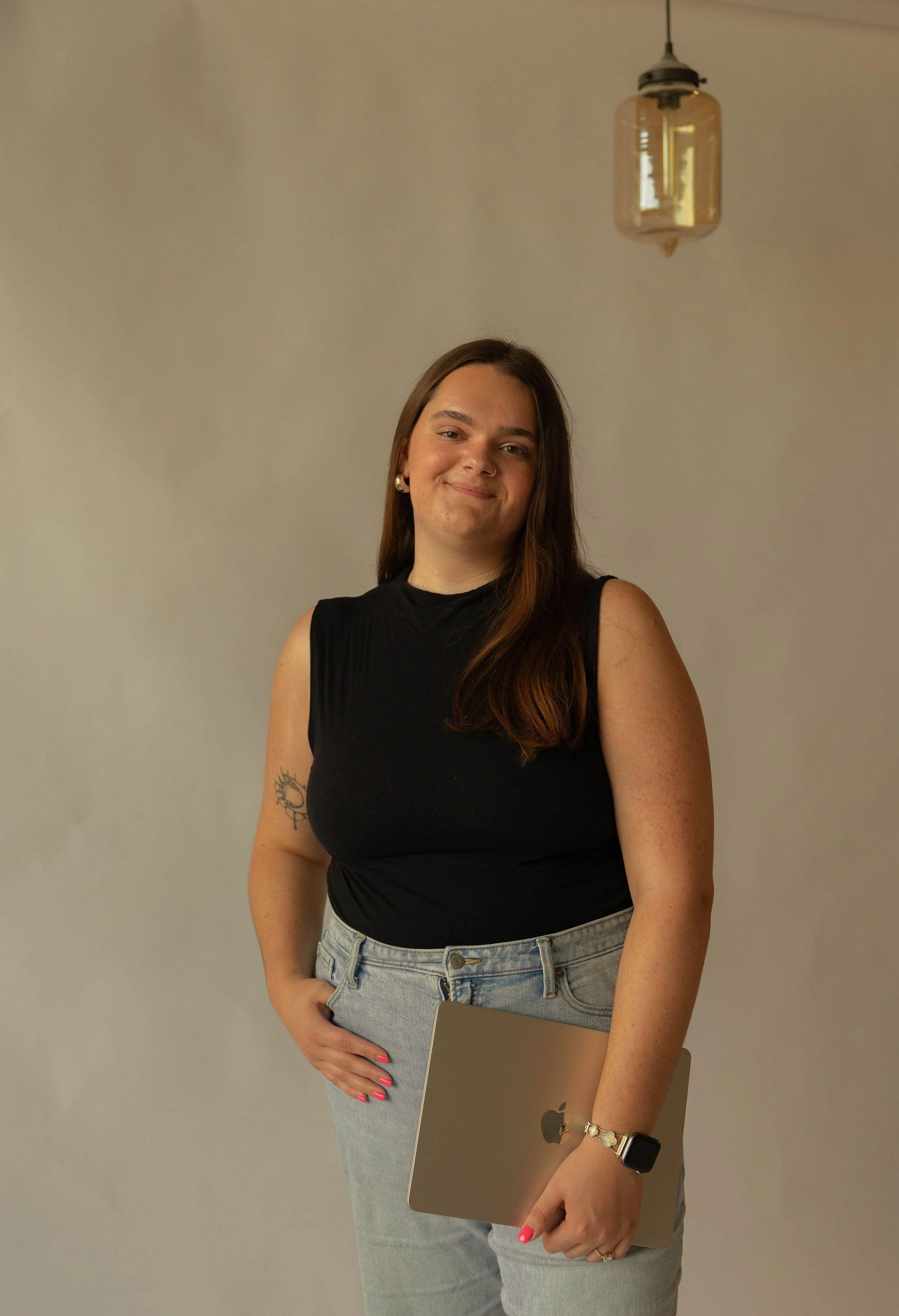 A smiling woman with long brown hair, wearing a black sleeveless top and light blue jeans, stands against a plain gray background holding a closed MacBook laptop, with a smartwatch and a bracelet on her wrist.
