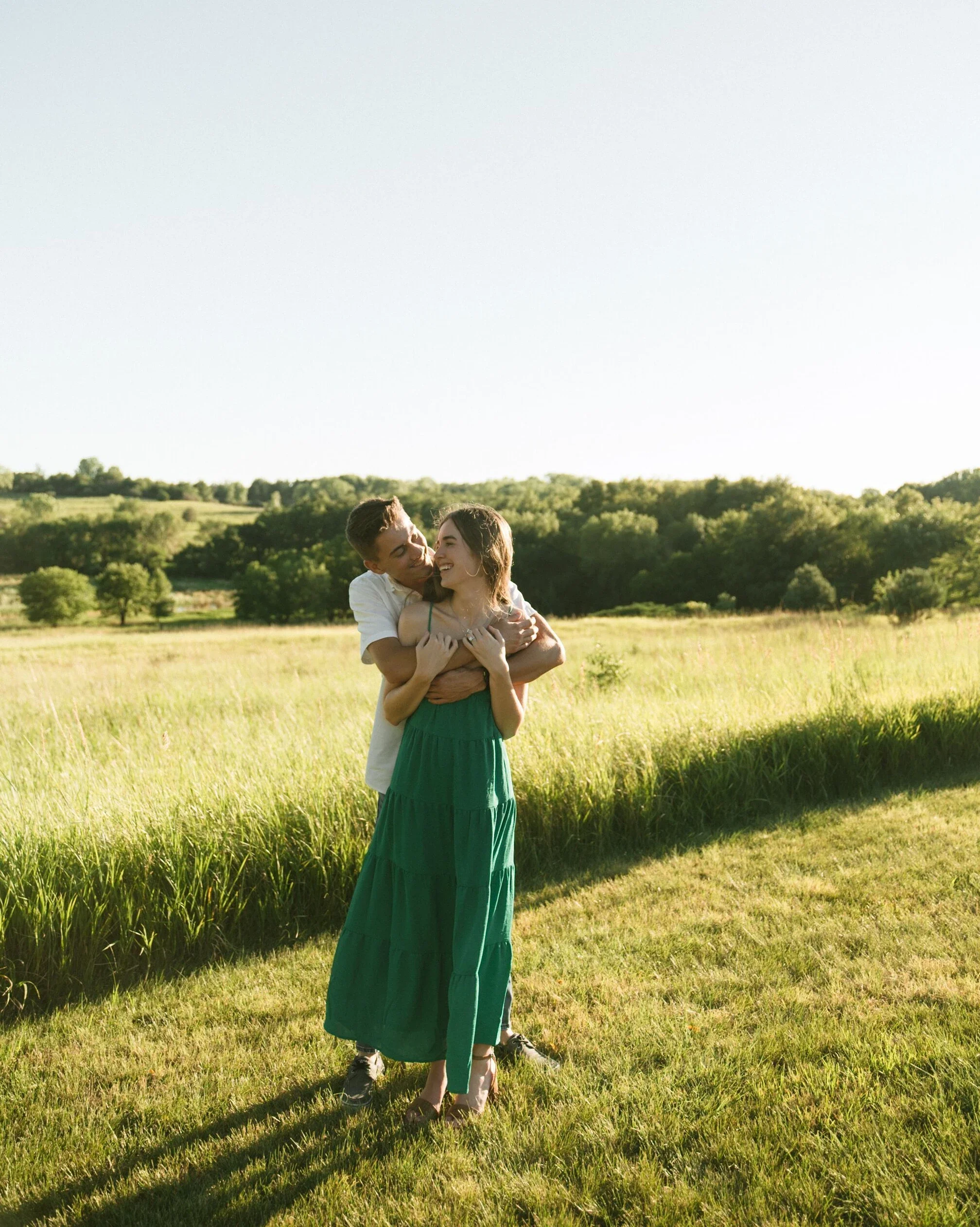 A young couple hugging each other in a grassy field with trees in the background on a sunny day.