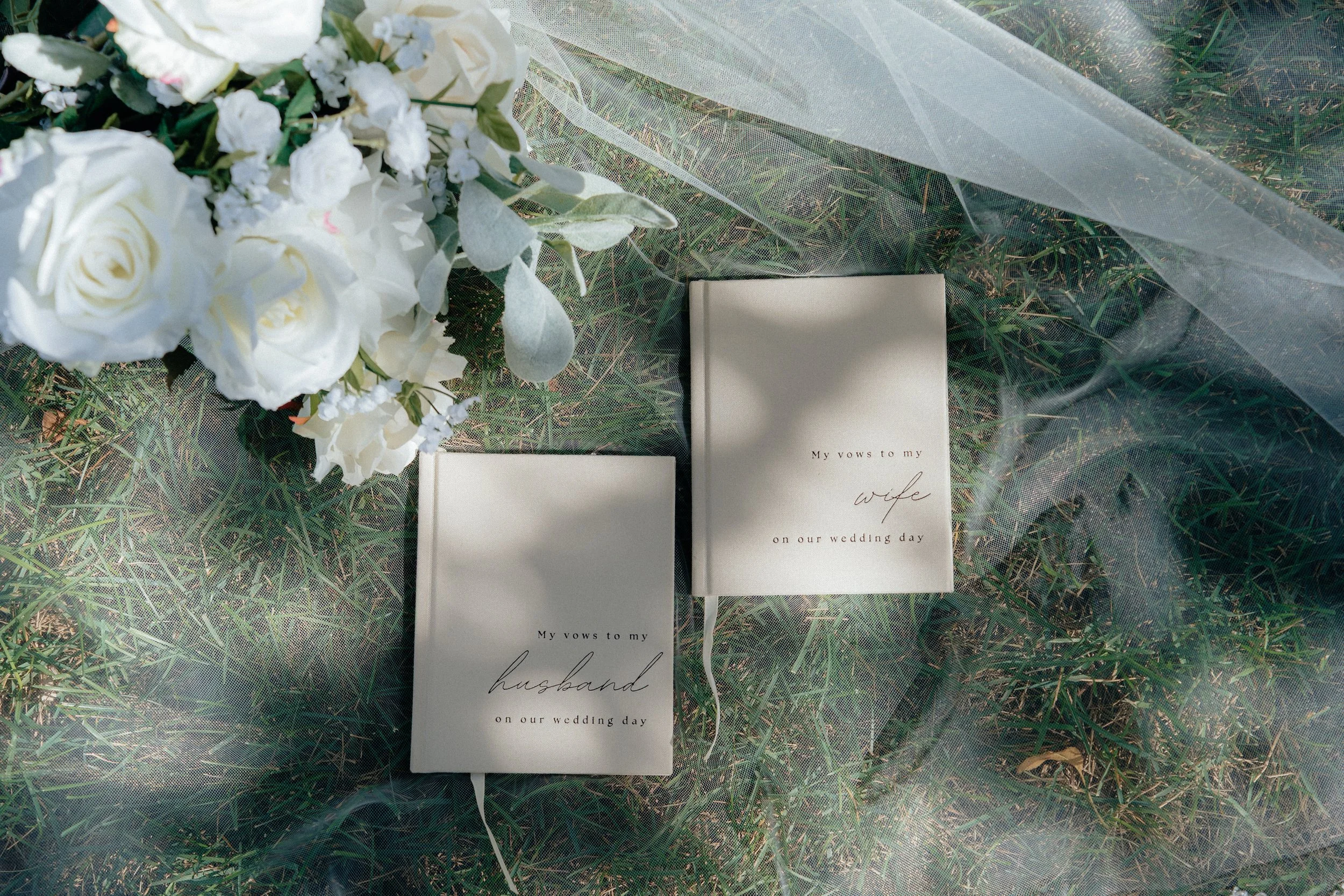 A wedding bouquet of white flowers resting on grass, next to two wedding vows booklets with the words 'My vows to my wife/husband on our wedding day' on the covers.