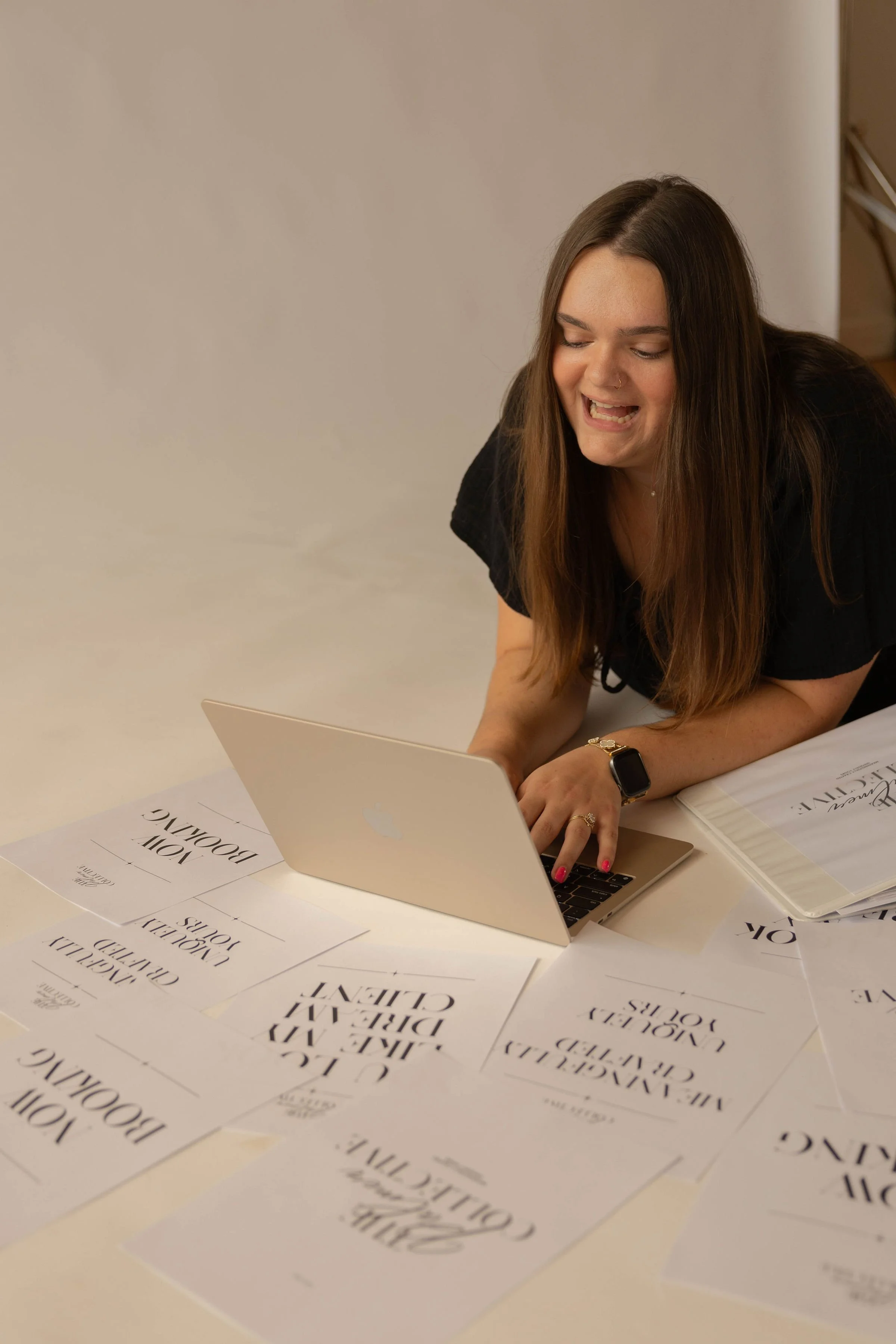 A woman with long brown hair, wearing a black shirt and smartwatch, sitting at a table with a laptop and several printed sheets with the text "MOVE ON BY WORKING CREATIVELY" and "JOY OF BLOOMING" scattered around.