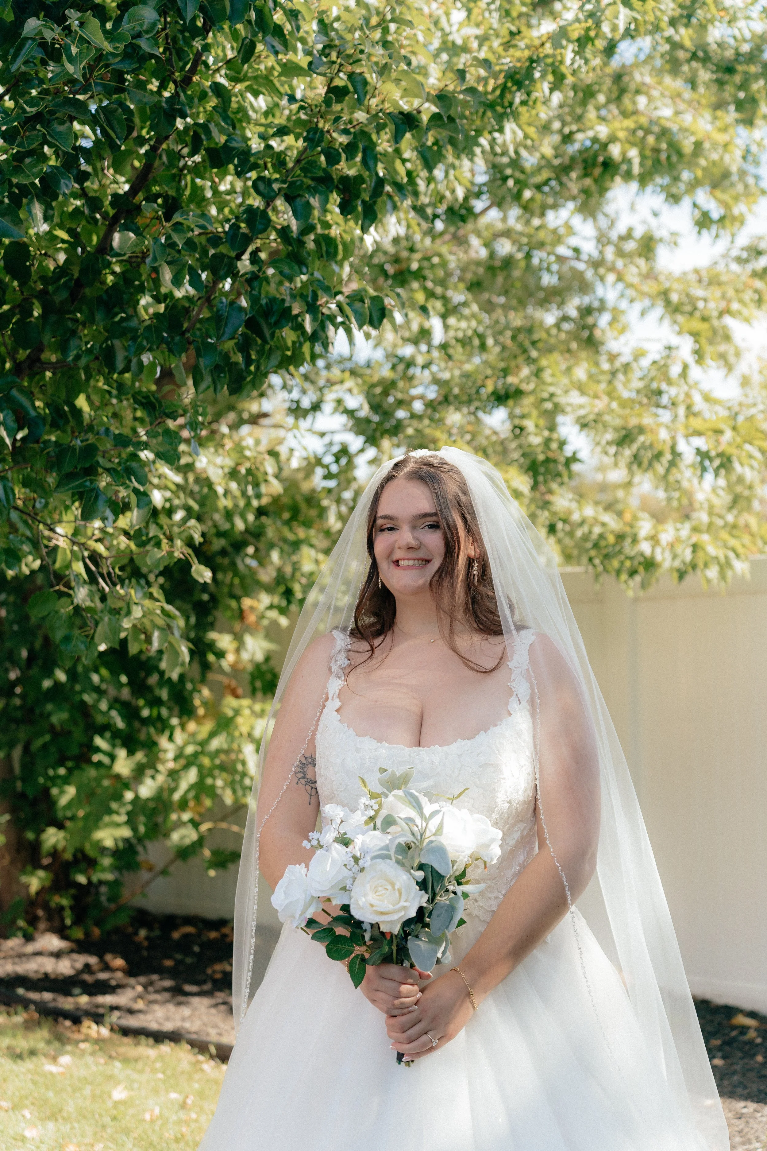 A bride in a wedding dress and veil holding a bouquet of white and green flowers, standing outdoors with green foliage and trees in the background.
