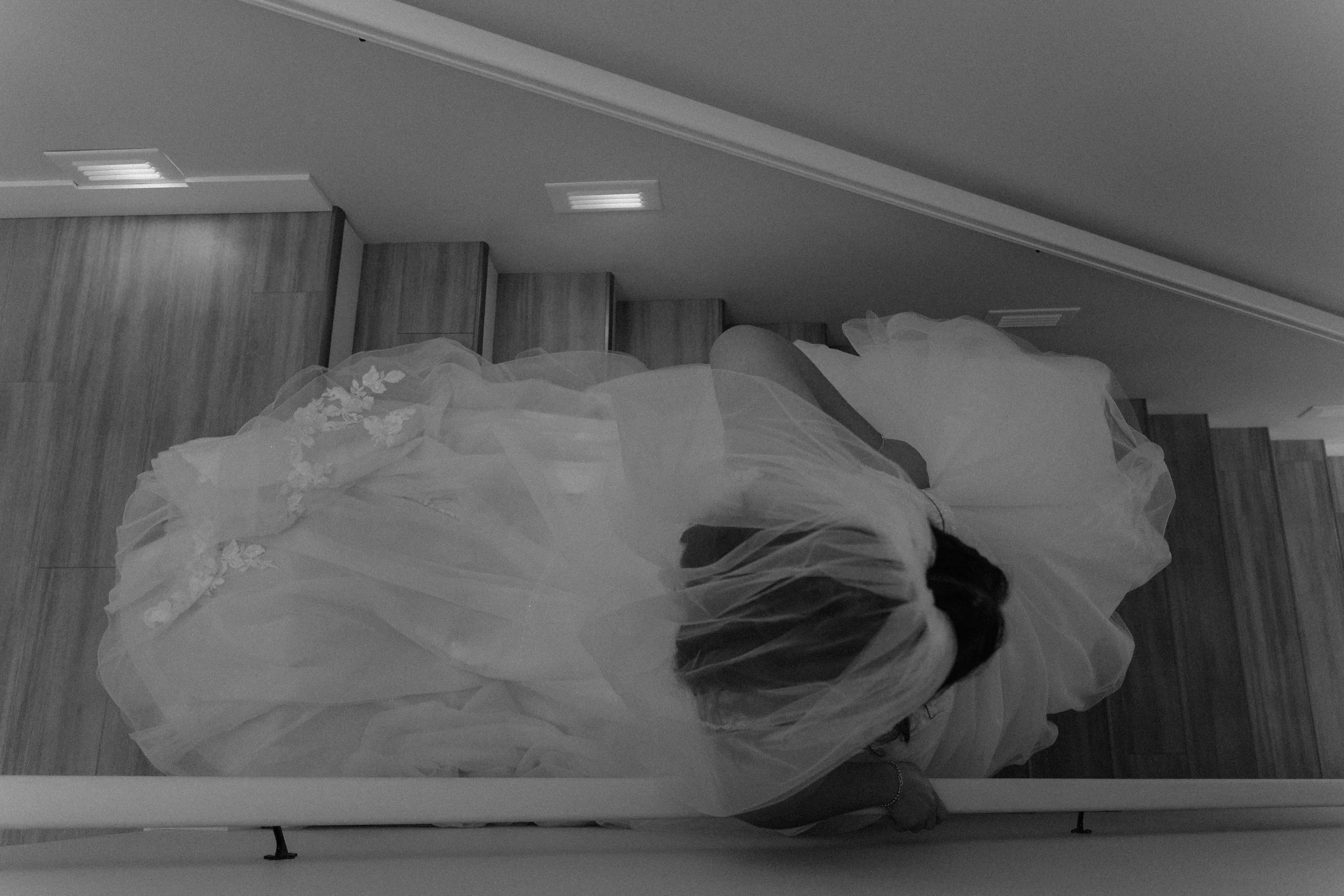 A person wearing a wedding dress with lace details, lying on a bed with a wooden headboard, in a room with ceiling lights and wooden flooring.