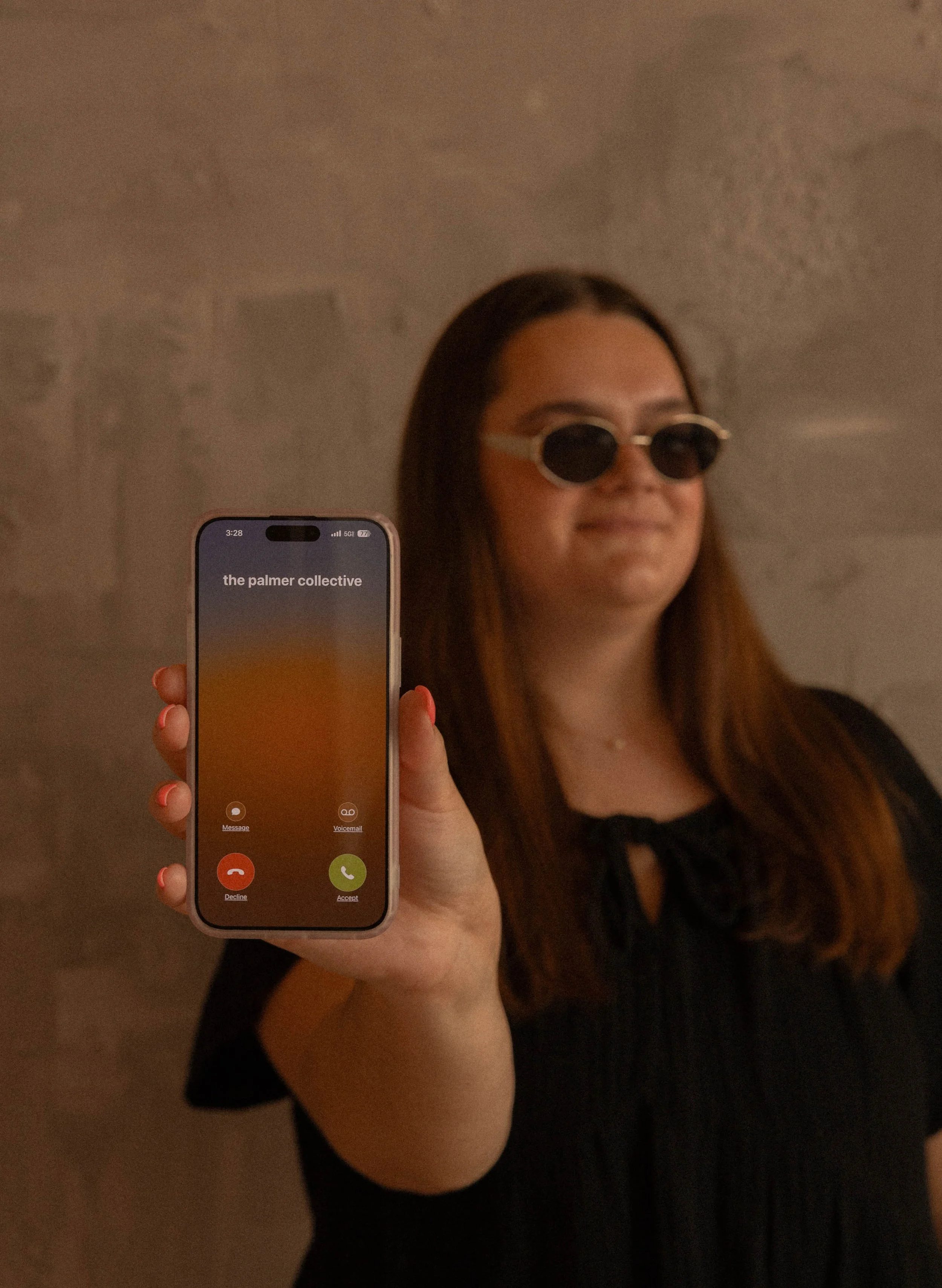 A woman wearing sunglasses is holding a smartphone displaying an incoming call screen that reads 'the palmer collective'.