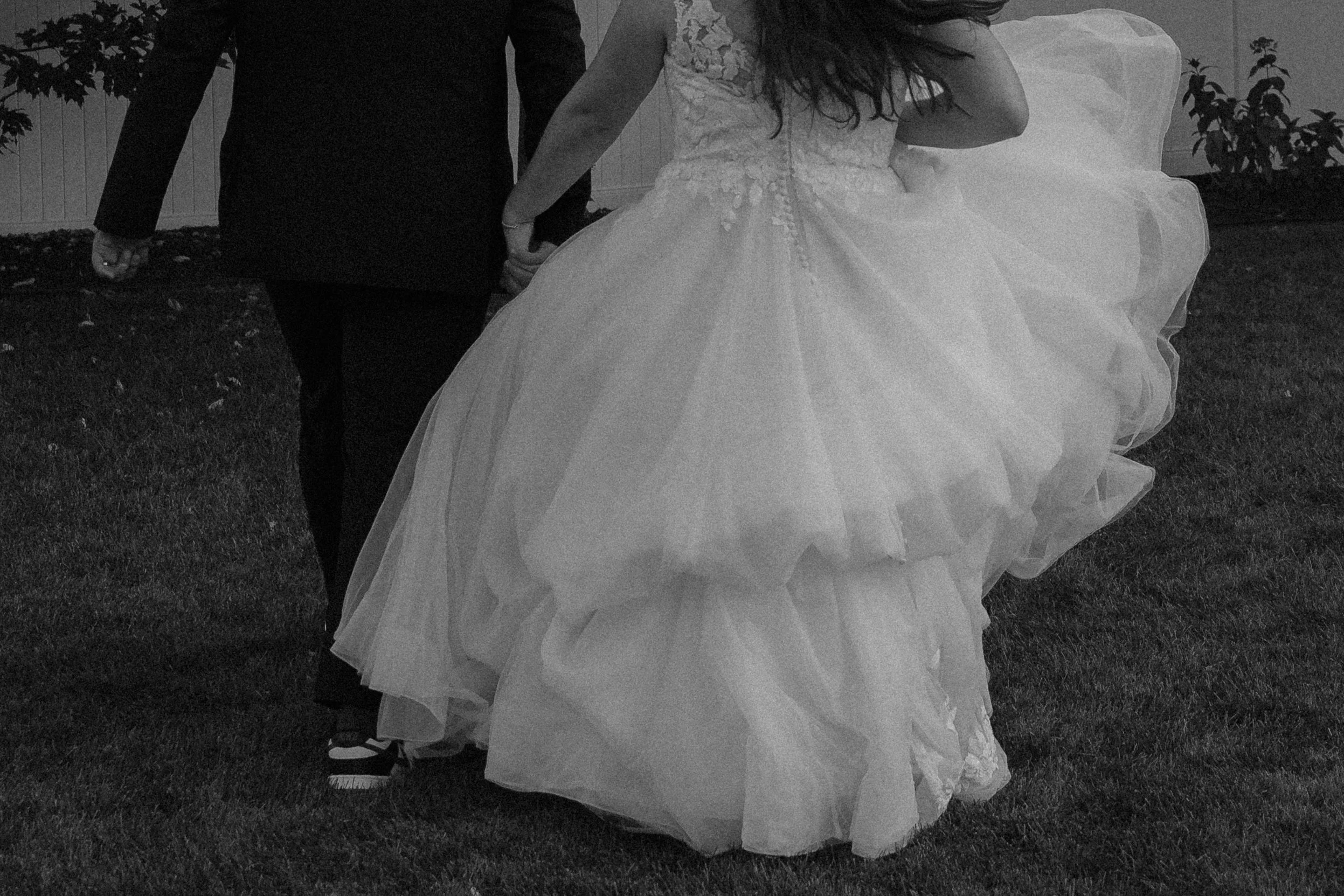 A bride in a wedding dress holding hands with a groom in a suit, standing outdoors on grass with trees and a white fence in the background.