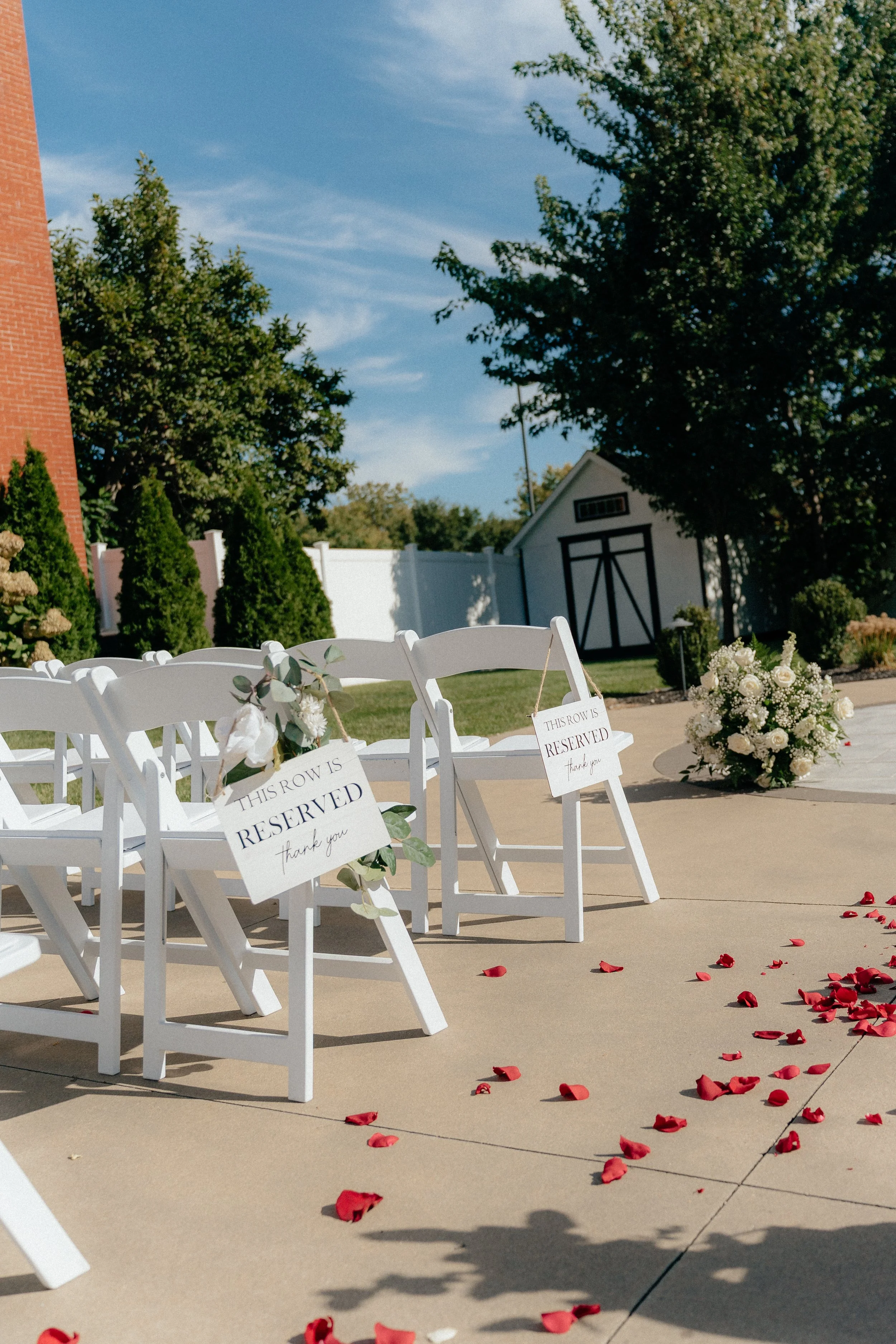 Outdoor wedding seating with white chairs, flower arrangements, and scattered red rose petals on the ground, with trees, a white fence, and a small garden shed in the background.
