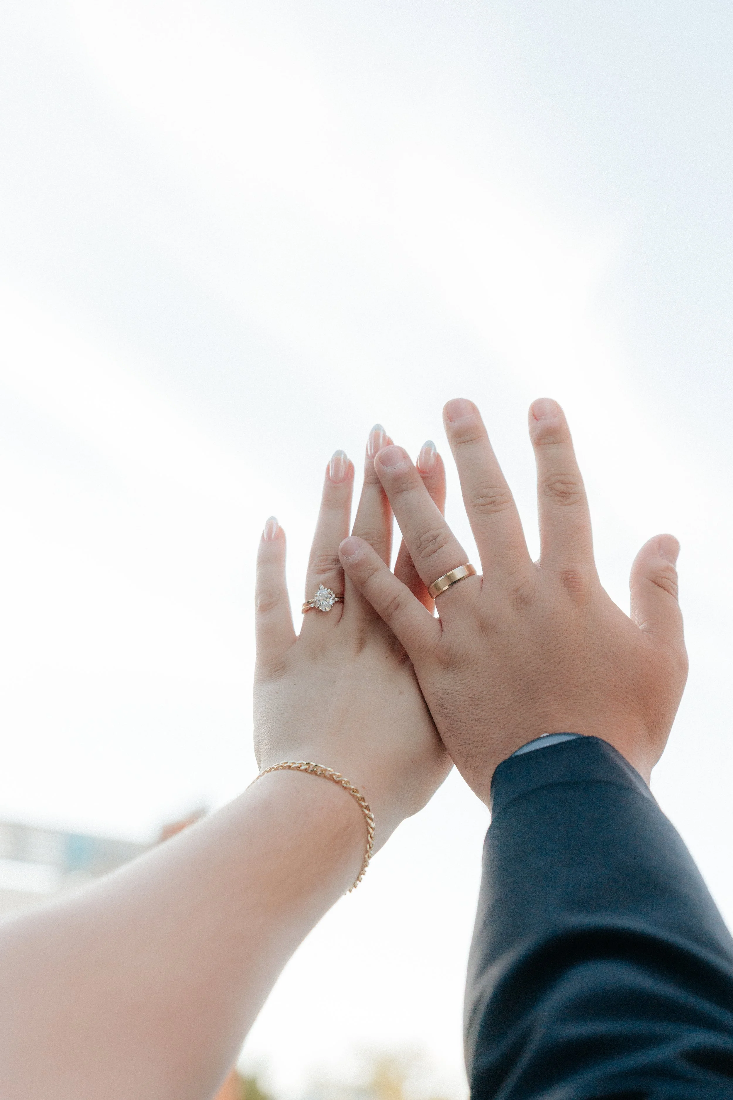 Close-up of two hands, one of a woman with light skin, wearing an engagement ring with a large diamond, and the other of a man with darker skin, wearing a wedding band, touching or holding hands against a cloudy sky background.
