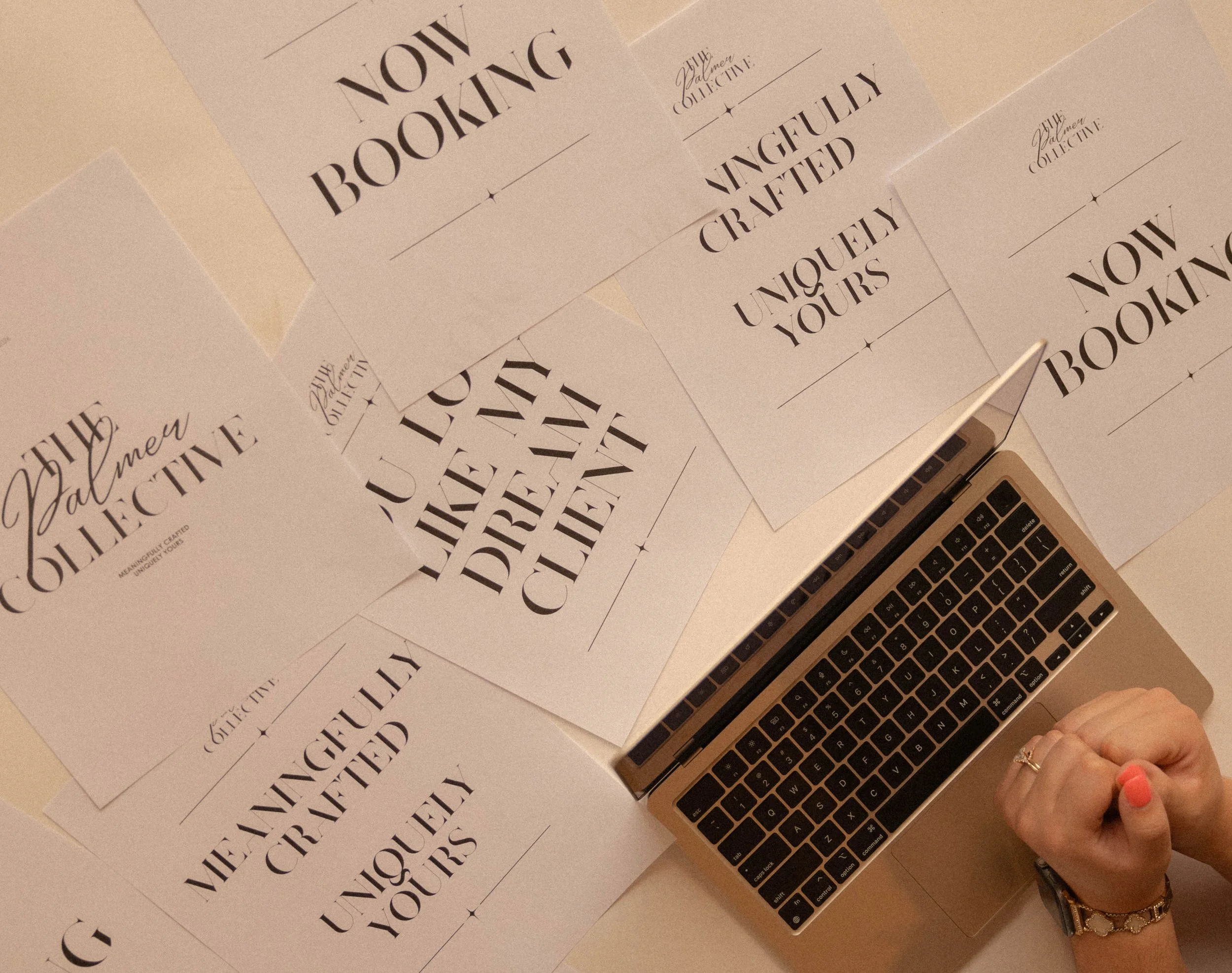 A person's hand with pink nail polish and a ring on the finger, using a laptop keyboard, surrounded by numerous printed sheets of paper with bold black text saying "NOW BOOKING," "VENGEFUL CRAFTED," "UNIQUE YOURS," and other designs.