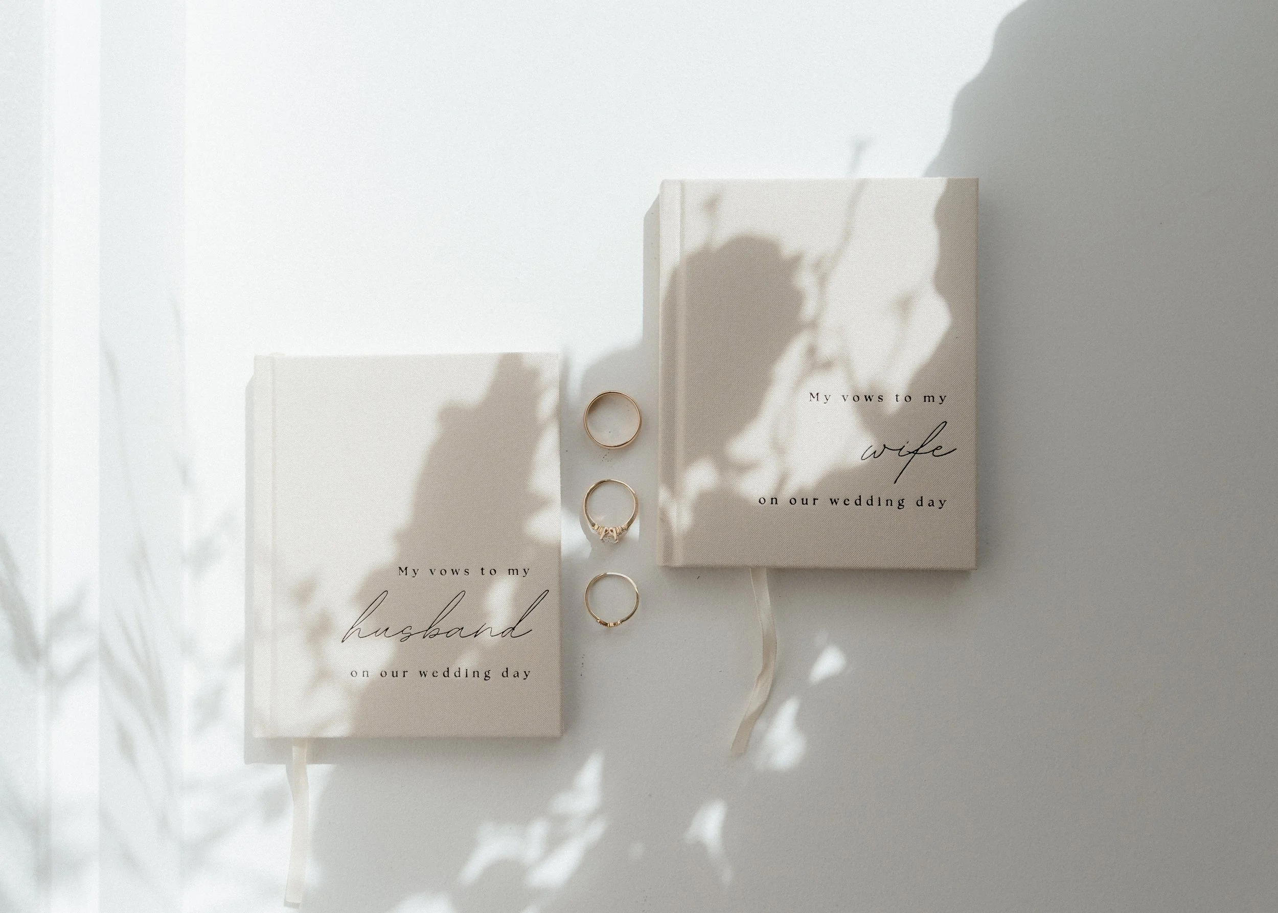 A white wedding vow book with the words "My vows to my wife" written on it, with three gold rings and a smaller vow booklet beside it, all casting shadows on a white surface.