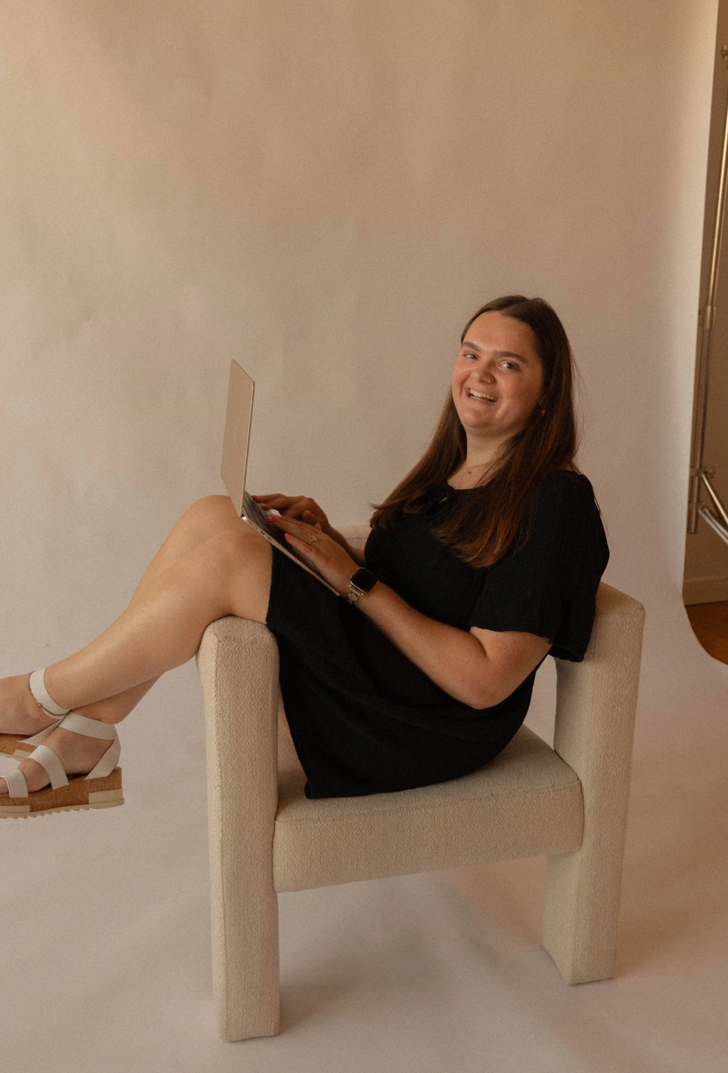 A woman with long brown hair, wearing a black dress, white sandals, and a smartwatch, is sitting on a beige armchair with her legs crossed. She is smiling and holding a laptop on her lap. The background features a plain beige wall and part of a door.