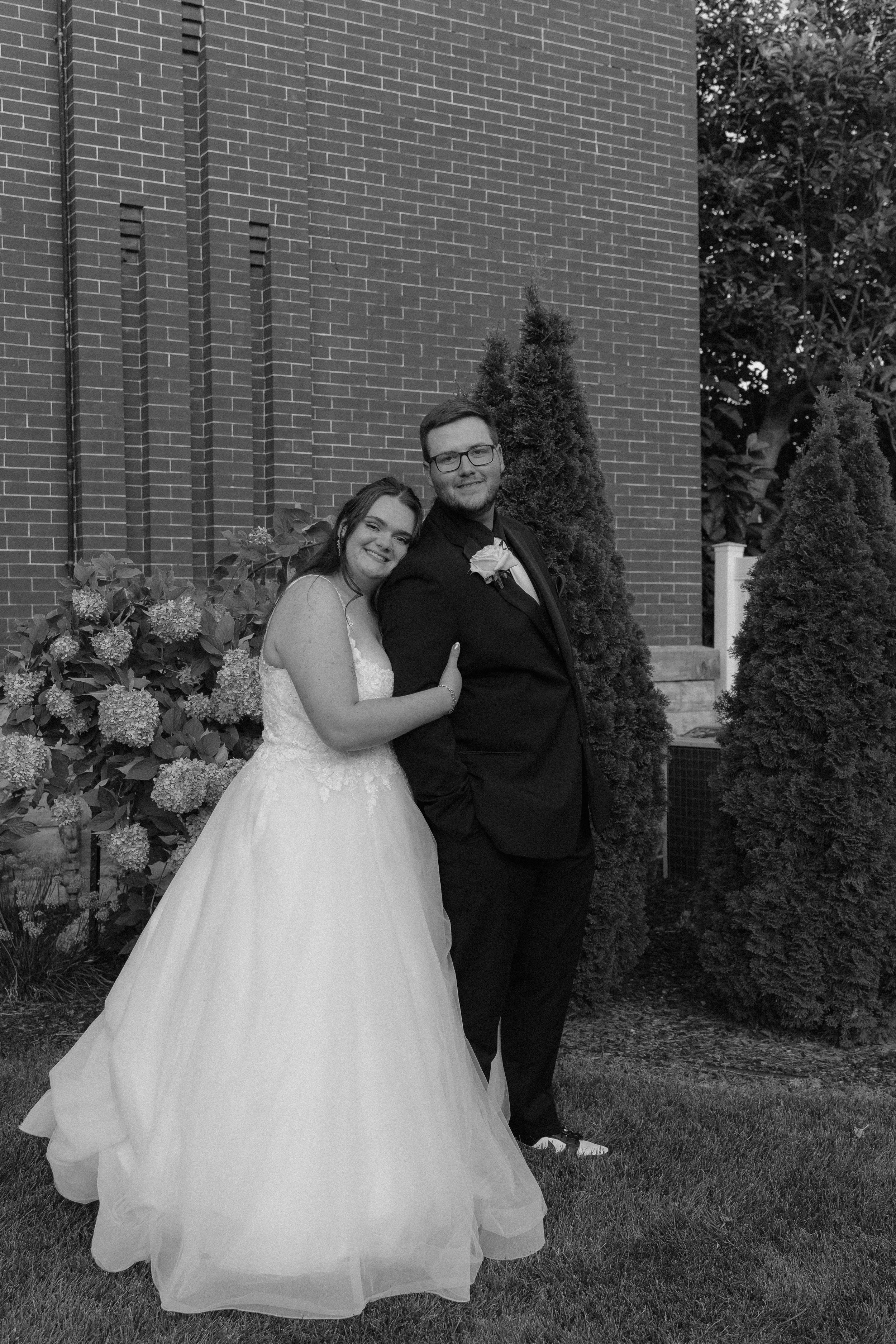 A bride and groom standing by a pond in a park with green trees in the background, smiling and embracing each other on their wedding day.
