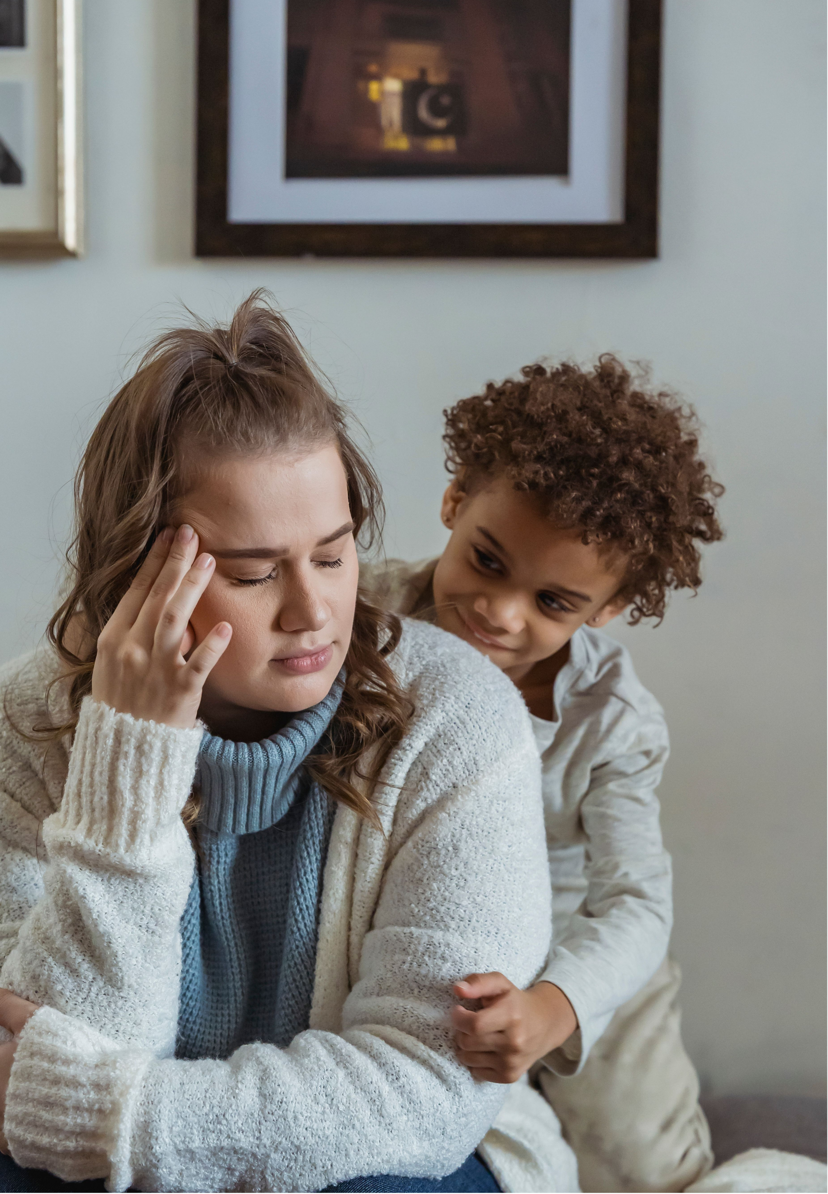 A woman with brown curly hair, wearing a white sweater and blue turtleneck, appears stressed with her hand on her forehead while a young boy with brown curly hair, wearing beige clothes, looks at her and smiles. They are sitting indoors against a wall with framed pictures.