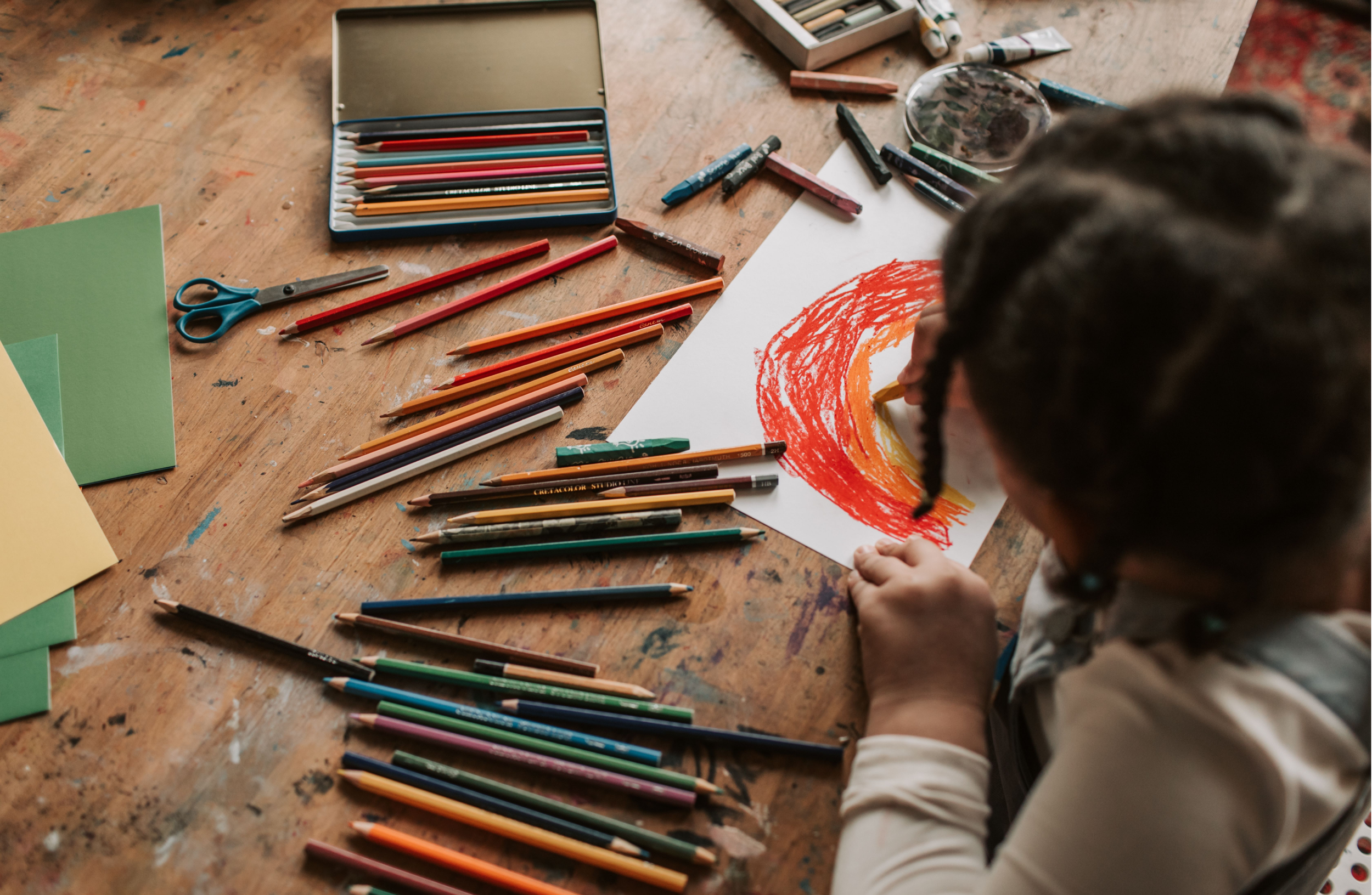 Child drawing with orange and yellow crayons on a large white sheet of paper, surrounded by various colored pencils and art supplies on a wooden table.