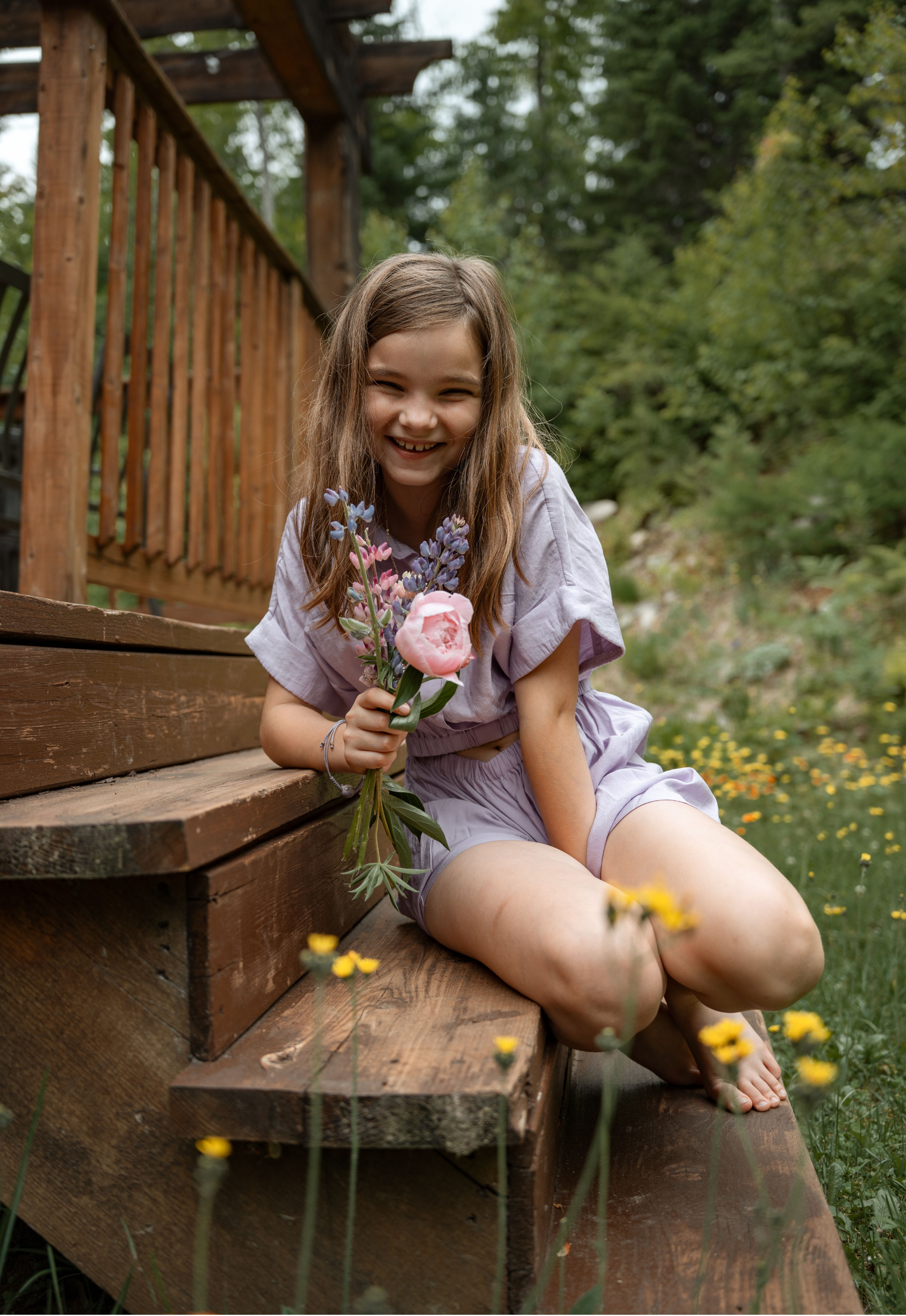 A young girl with long brown hair sitting on wooden outdoor stairs, holding a bouquet of flowers, smiling cheerfully, surrounded by green trees and yellow flowers.