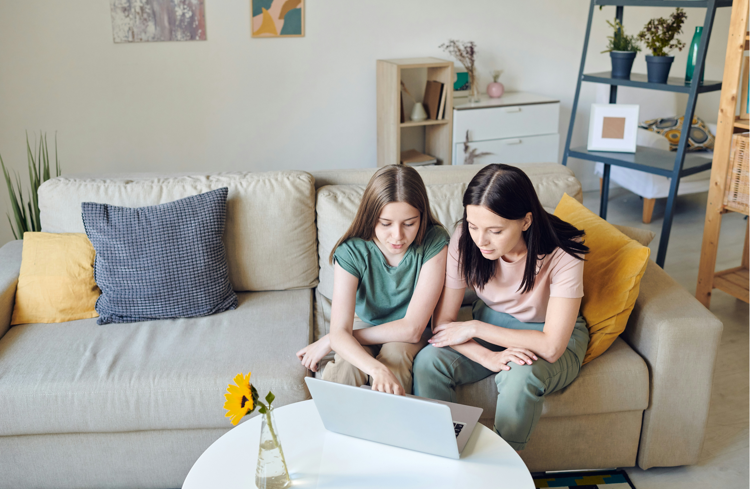 Two women sitting on a beige sofa in a living room, looking at a laptop. The sofa has yellow and blue pillows. There is a white table with a small vase containing a sunflower. In the background, there are art pieces on the wall, a bookshelf, and a shelf with plants.