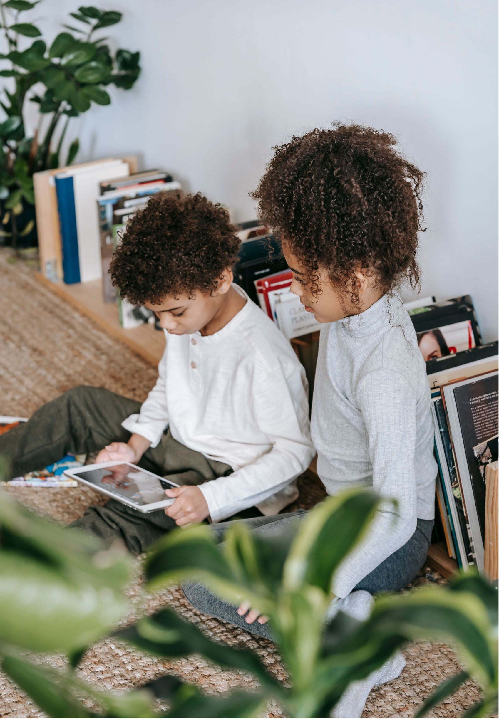 Two children with curly hair sitting on a woven rug inside, looking at a tablet device. One child is holding the tablet while the other stands beside, both focused on the screen. Books and magazines are visible in the background, and part of a green plant is in the foreground.