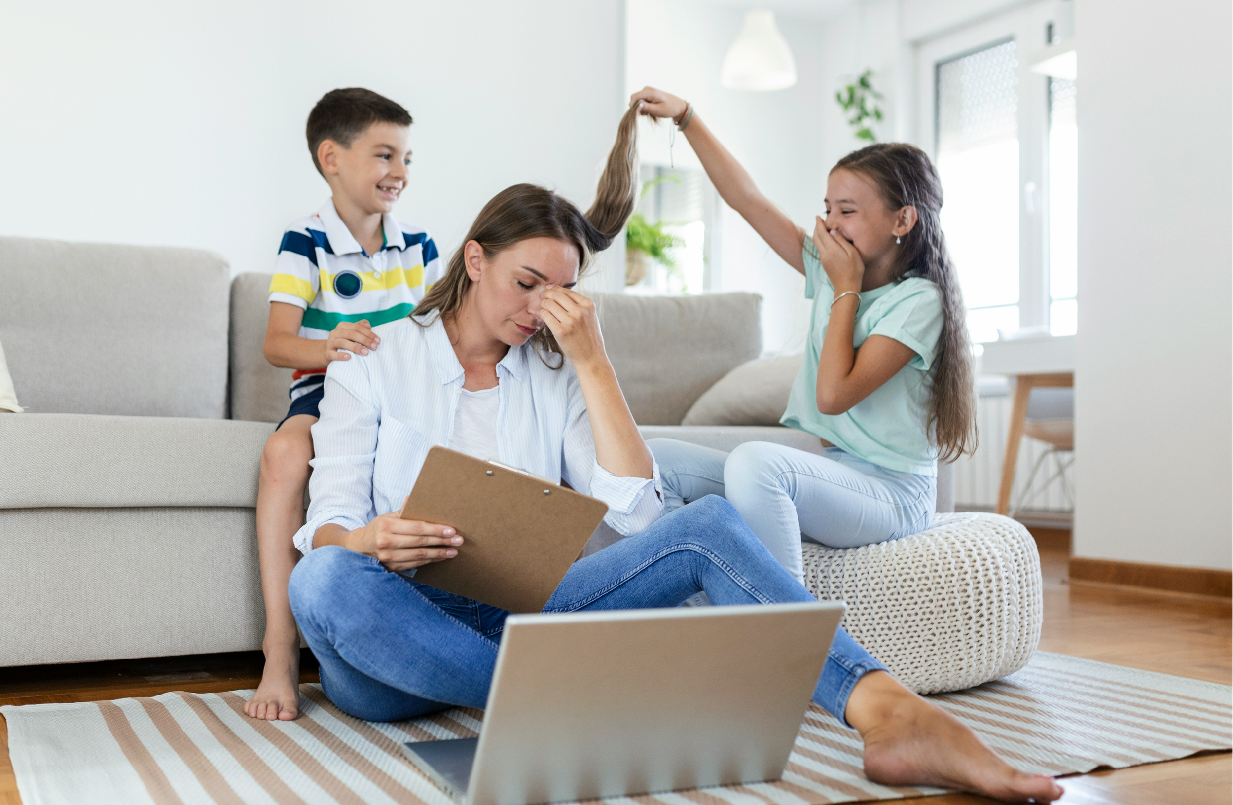 A woman sitting on the floor with her head in her hand, surrounded by two children. One child is holding her hair as if pulling or playing with it, and the other is smiling at them. There is a laptop on the floor in front of her.