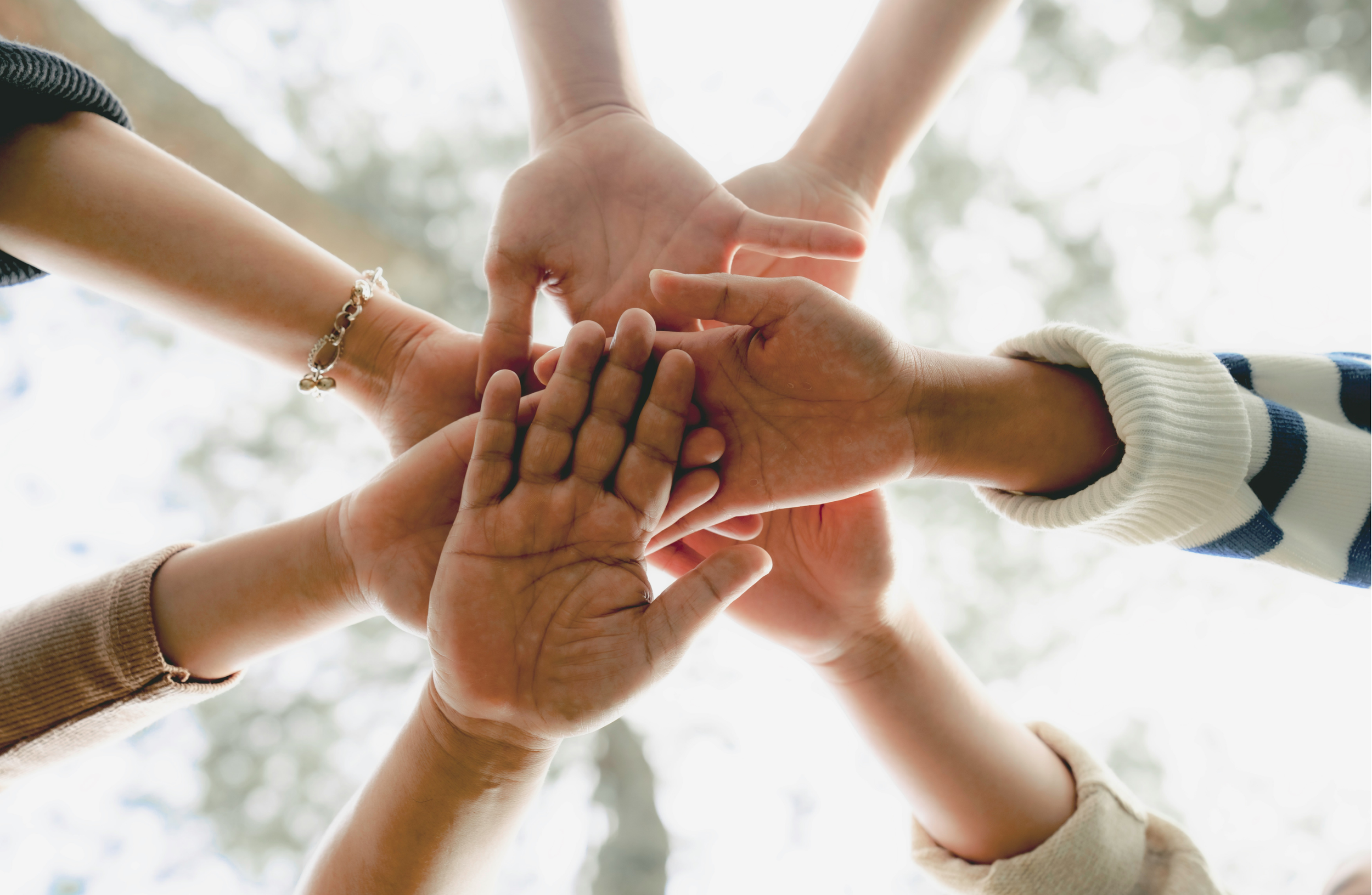 Multiple hands of diverse individuals placed together in the center, forming a circle outdoors with blurred trees in the background.