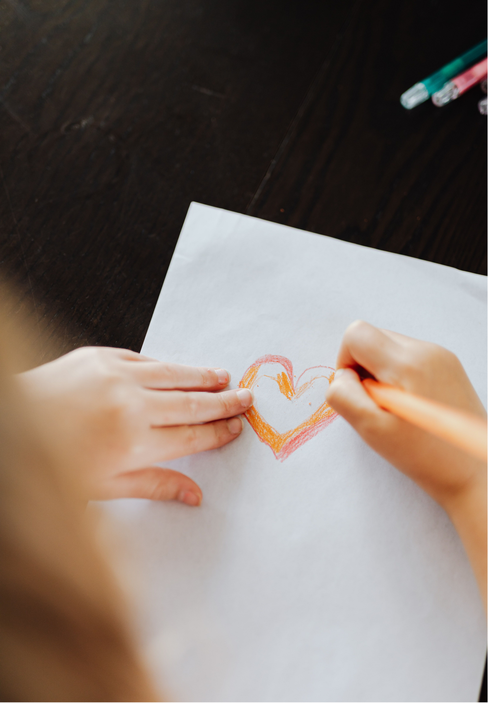 Child's hand coloring a heart on white paper with orange and red crayons, on a dark wooden table.