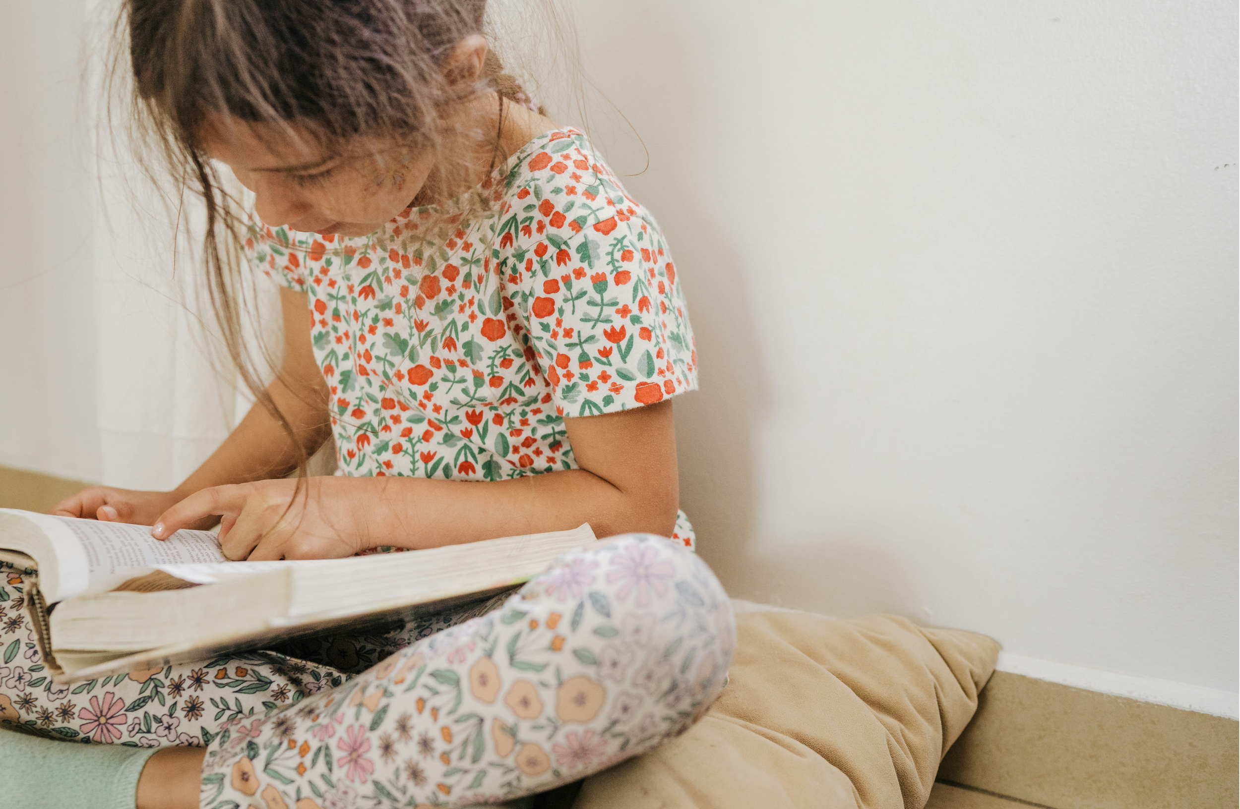 A young girl with long, curly hair is reading a book. She is sitting on a beige couch, wearing a floral dress with red, green, and white colors, and floral leggings.