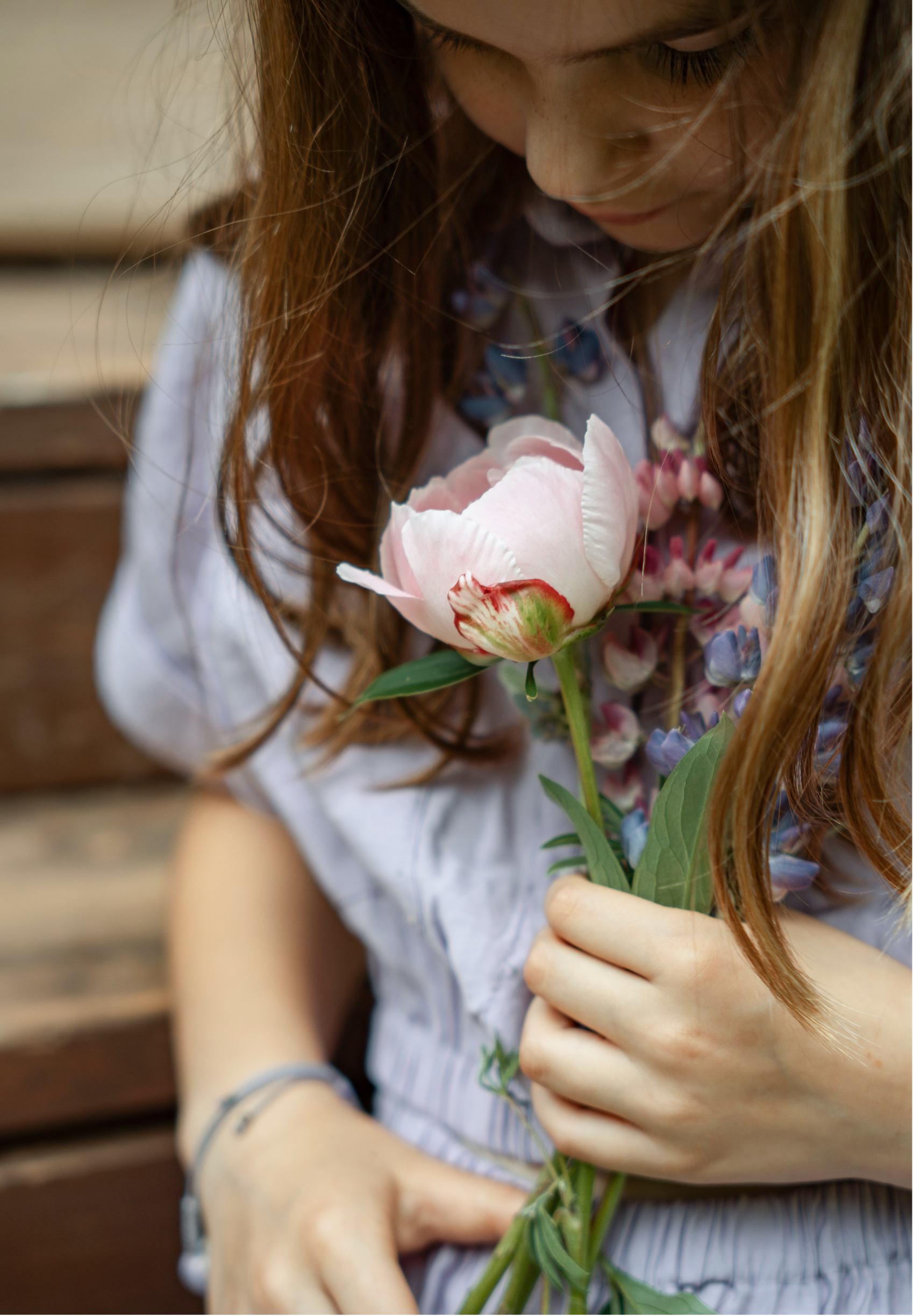 A young girl with long reddish hair holding a pink peony flower in front of her face, sitting outdoors on a wooden surface.