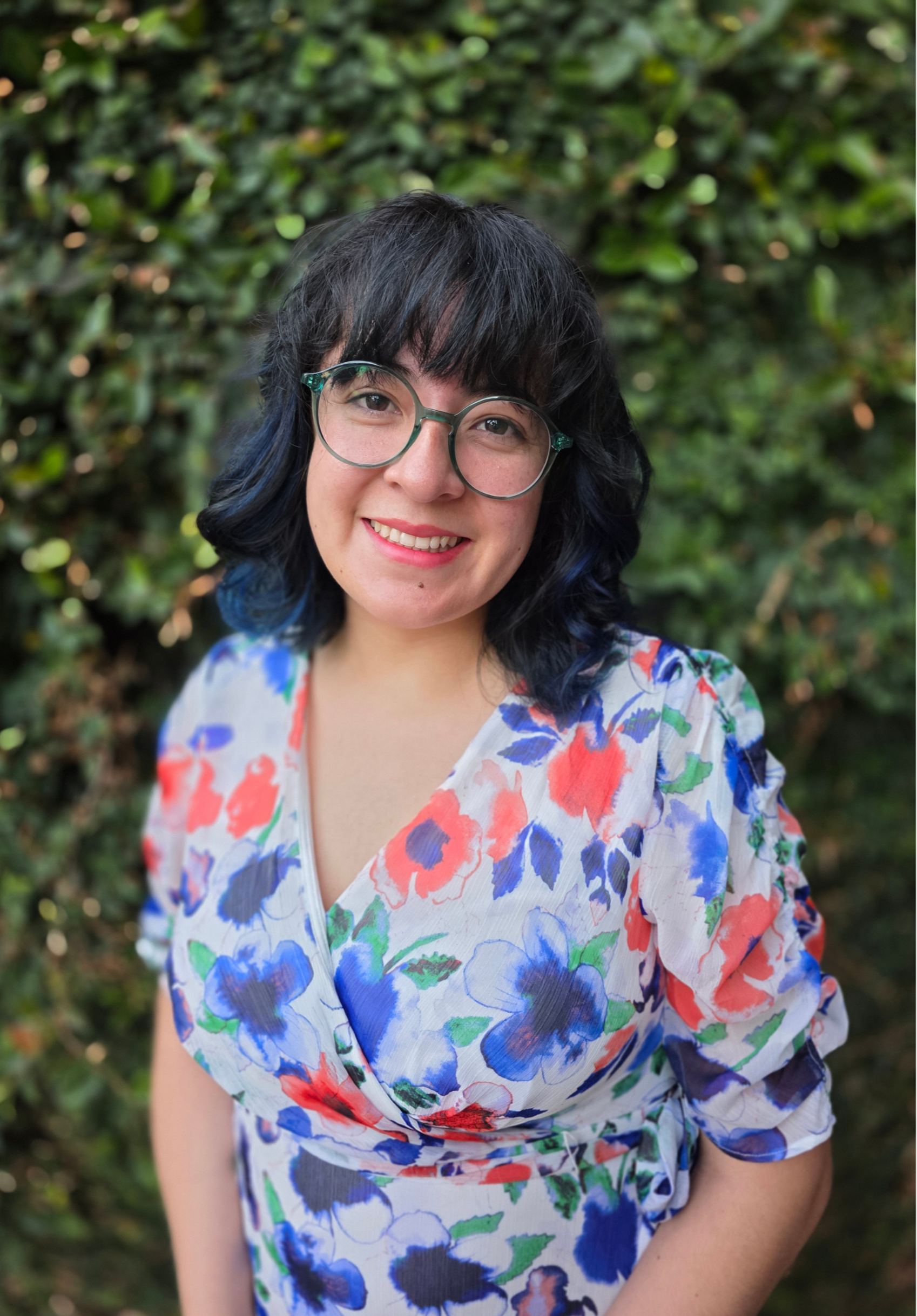 Anaymee Cifuentes LMFT, in California, with black hair, styled in loose waves with blue tips, wearing large round glasses and a colorful floral dress, standing outdoors in front of greenery and smiling.