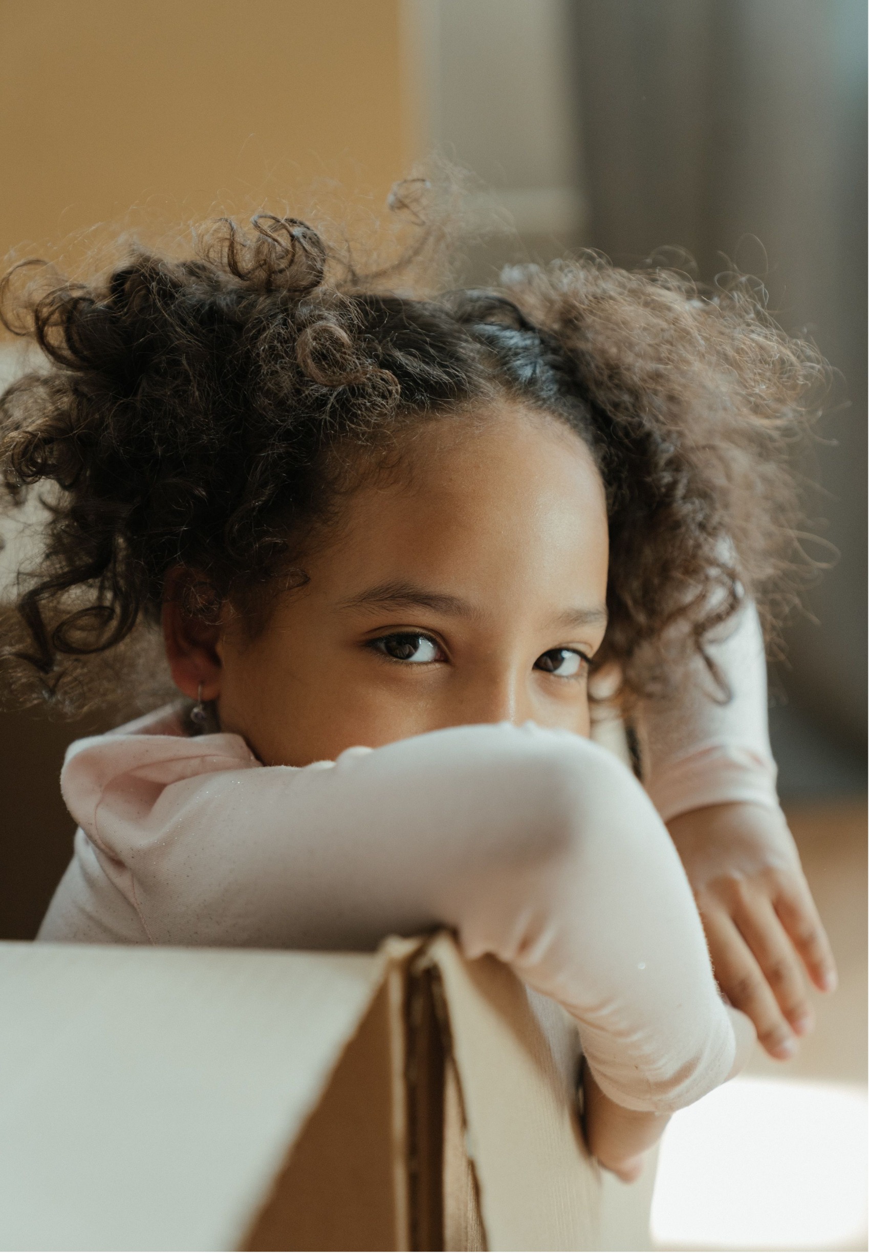 Close-up of a young girl with curly hair resting her head on her crossed arms, looking at the camera.
