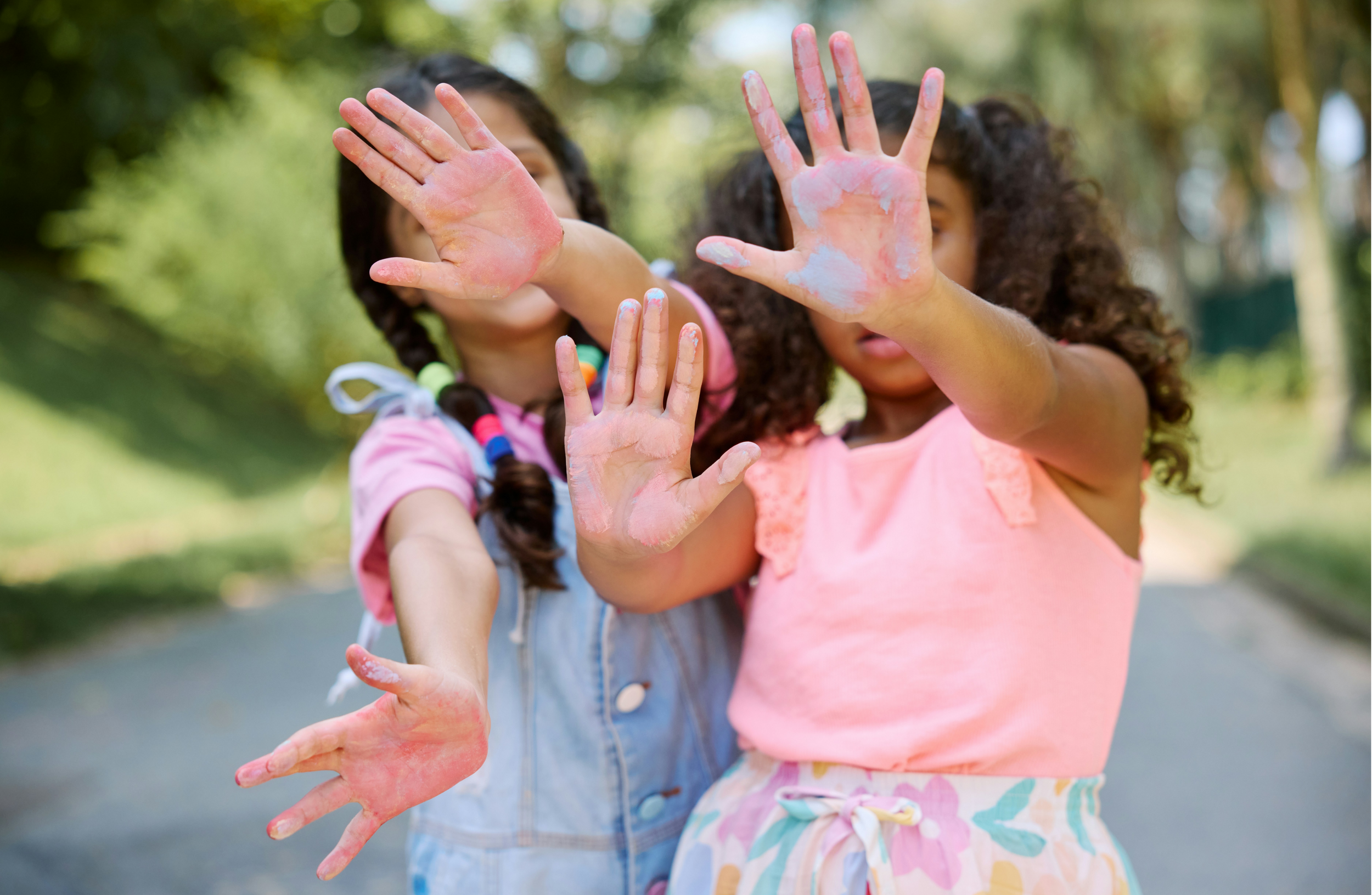 Two girls with curly hair showing their painted hands with pink and white paint outdoors.