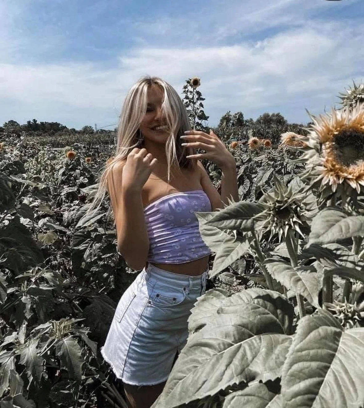 A woman with blonde hair in a purple strapless top and white shorts standing in a sunflower field, smiling and touching her hair.