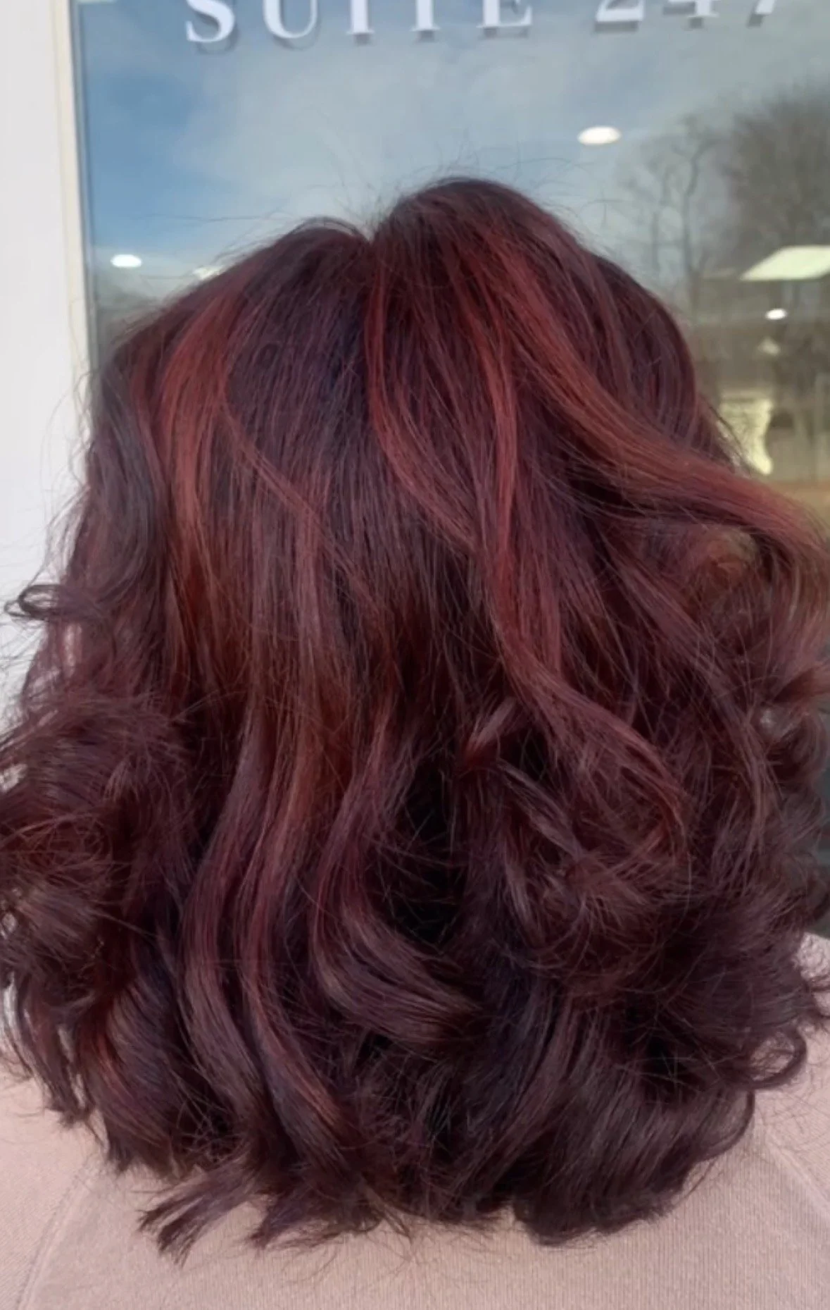 A person's head with red and burgundy styled curly hair, viewed from the back, in front of a glass door with the partial text 'SOUTH 24' visible.