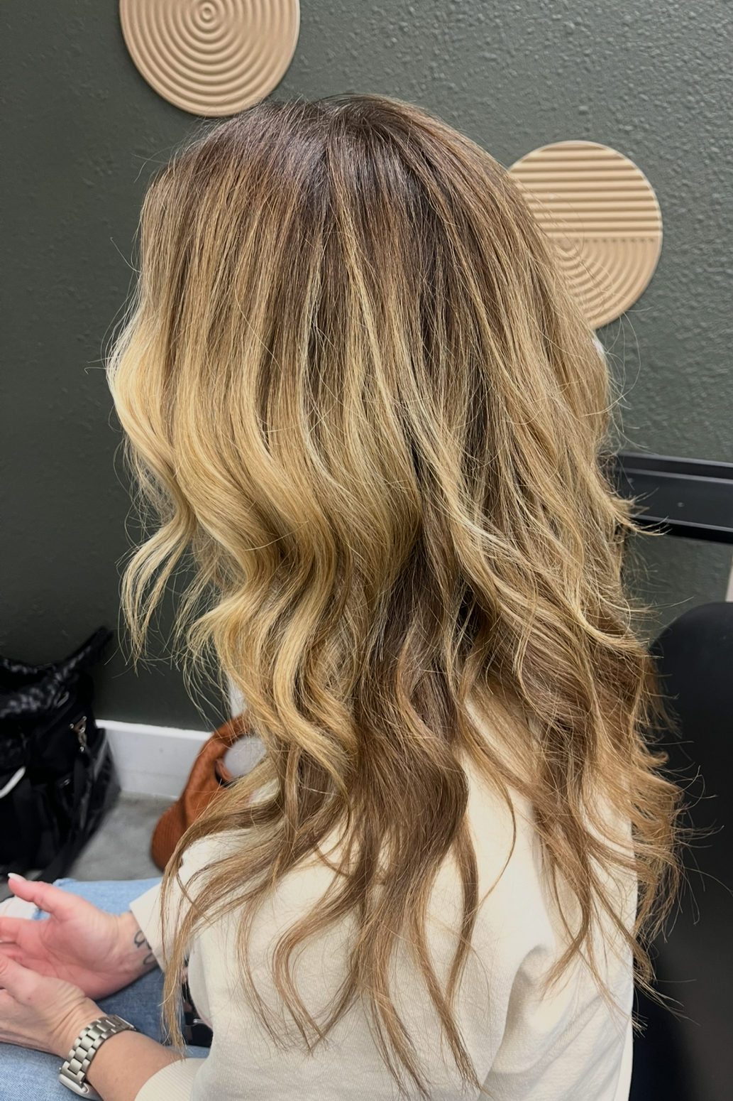 Woman with wavy blonde hair in a salon, sitting with her back to the camera.