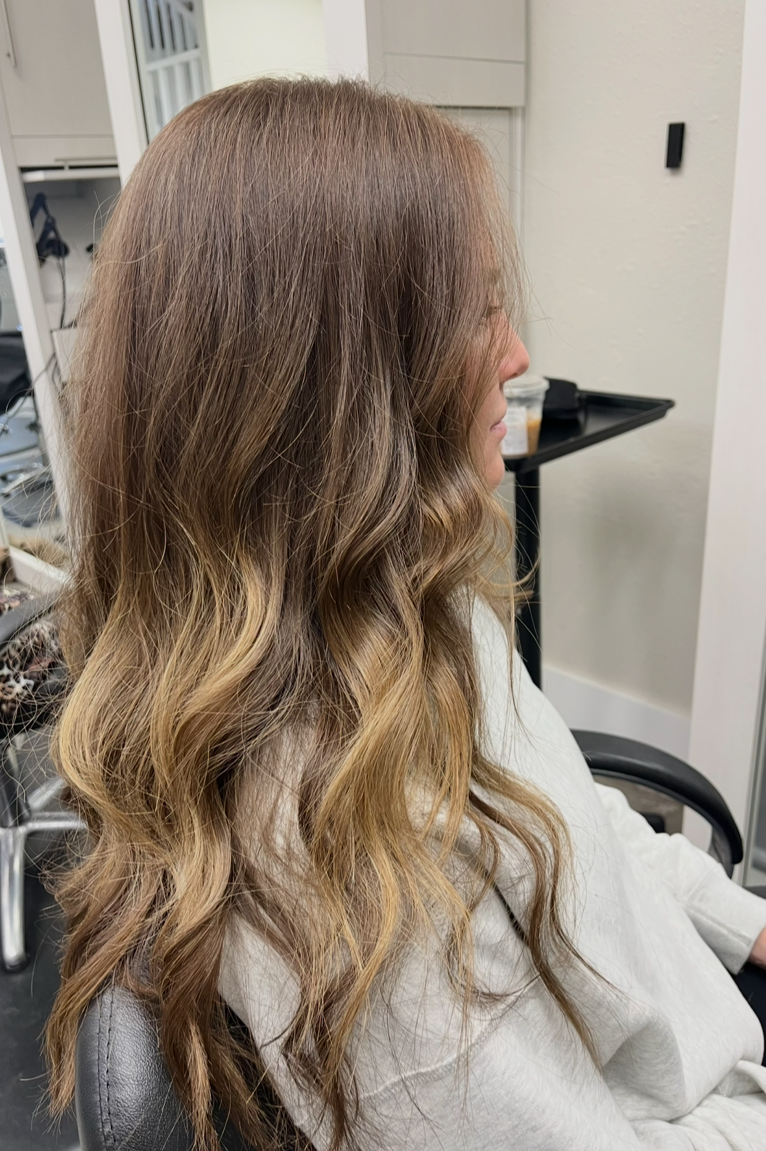 Side view of a woman with long, wavy, ombre hair in shades of brown and blonde, sitting in a salon chair.