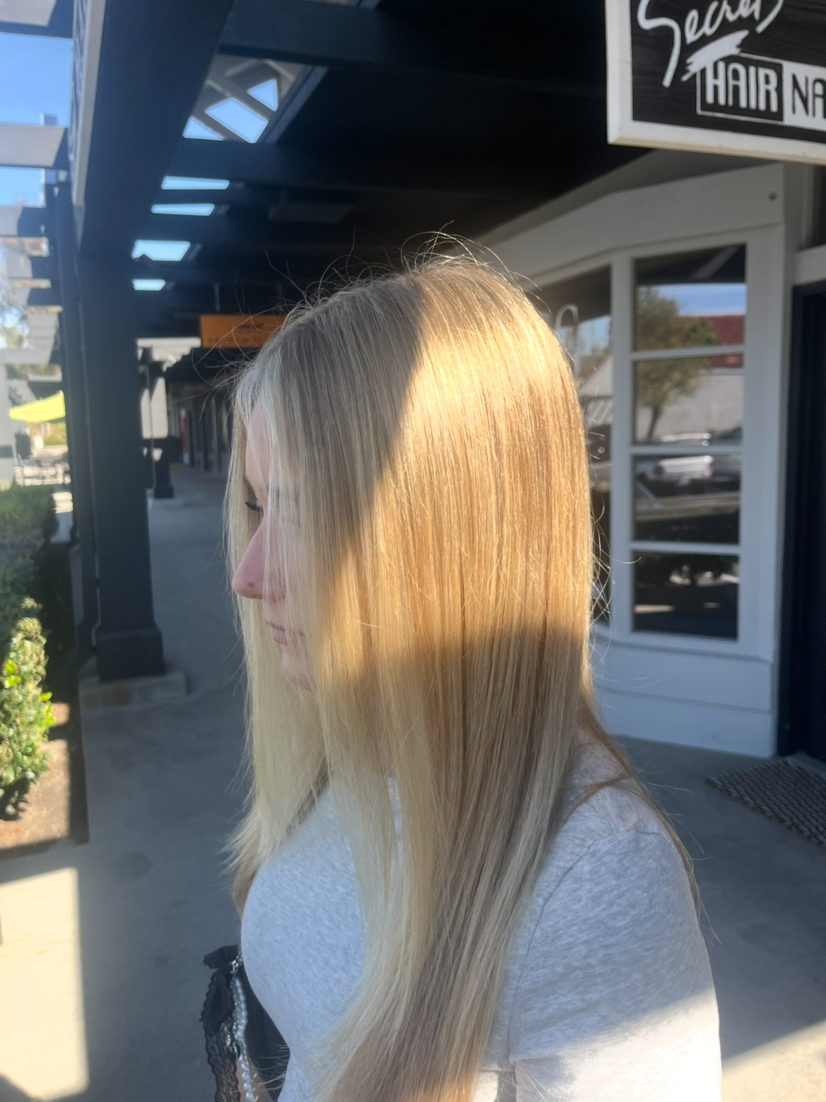 Side view of a woman with long blonde hair outside, partially shadowed, with storefront and signs in the background.