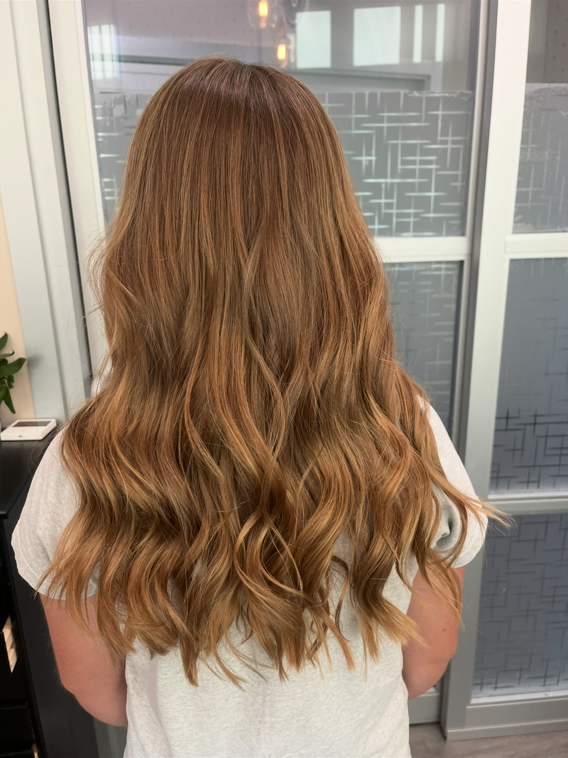 Back view of a woman with long, wavy, auburn hair standing indoors near glass doors.