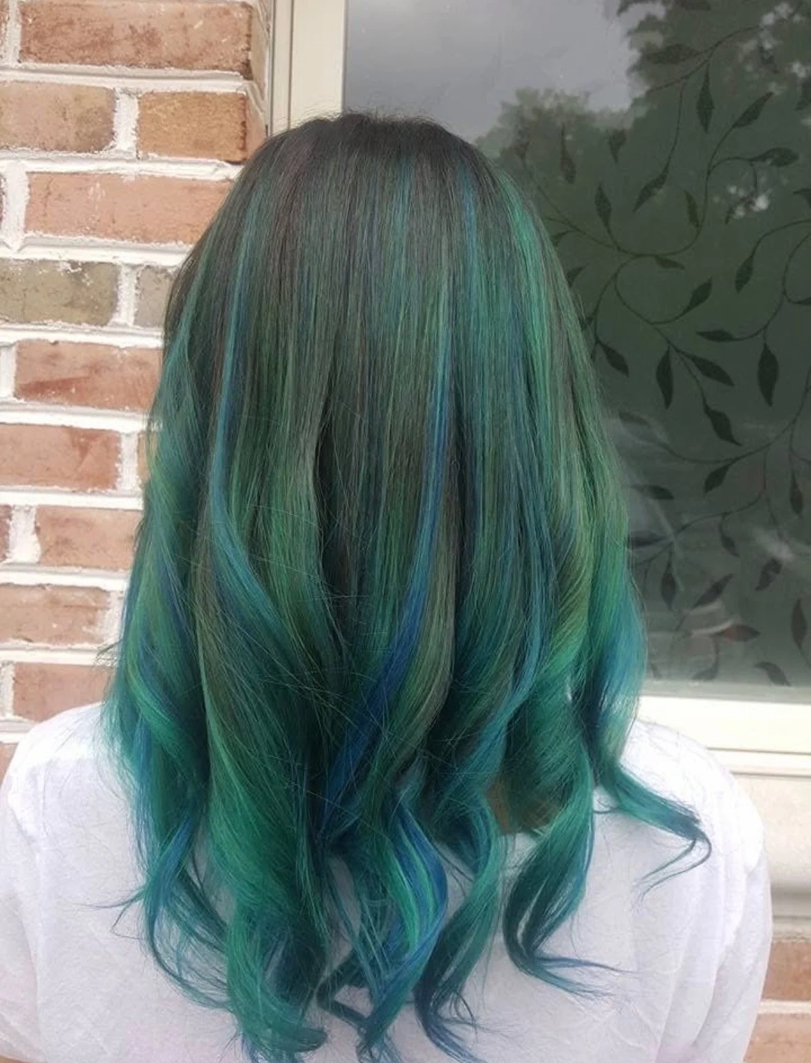 Back view of a person with long, wavy hair dyed in shades of blue, green, and teal, standing outdoors near a brick wall and window.