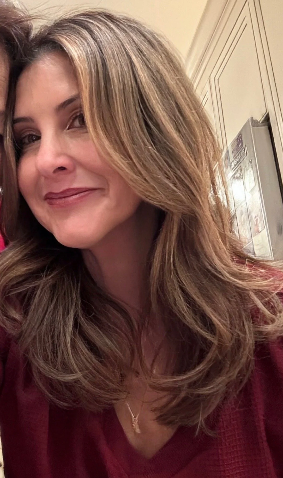 A woman with long, wavy, caramel-colored hair, wearing a red sweater and a gold necklace, smiling, indoors with beige walls and a wooden cabinet in the background.